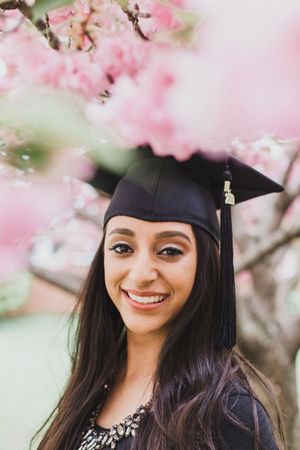 Graduate in cap and gown smiling outdoors with pink cherry blossoms in the background.