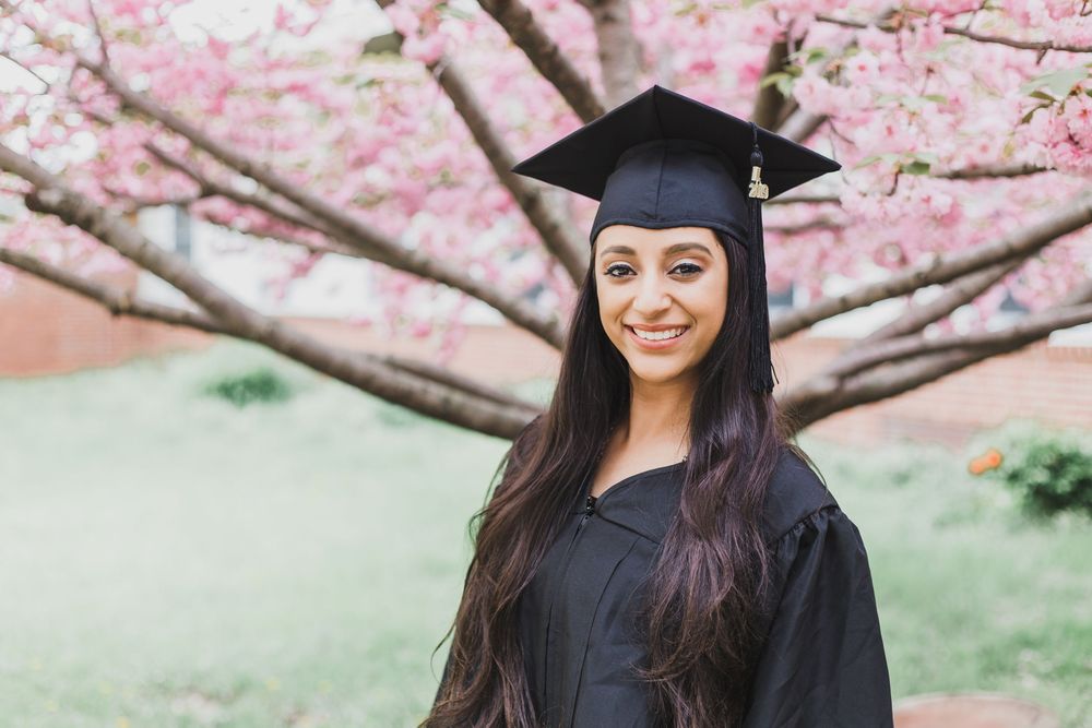 Graduate in cap and gown smiling under blooming pink cherry blossom trees during spring graduation photoshoot.