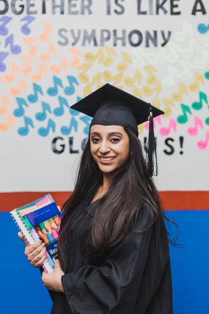 Graduate in black cap and gown holding textbooks poses against colorful wall with symphony-themed decorations.