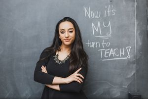 A confident educator stands with arms crossed in front of a chalkboard with text reading 'Now it's my turn to teach'.