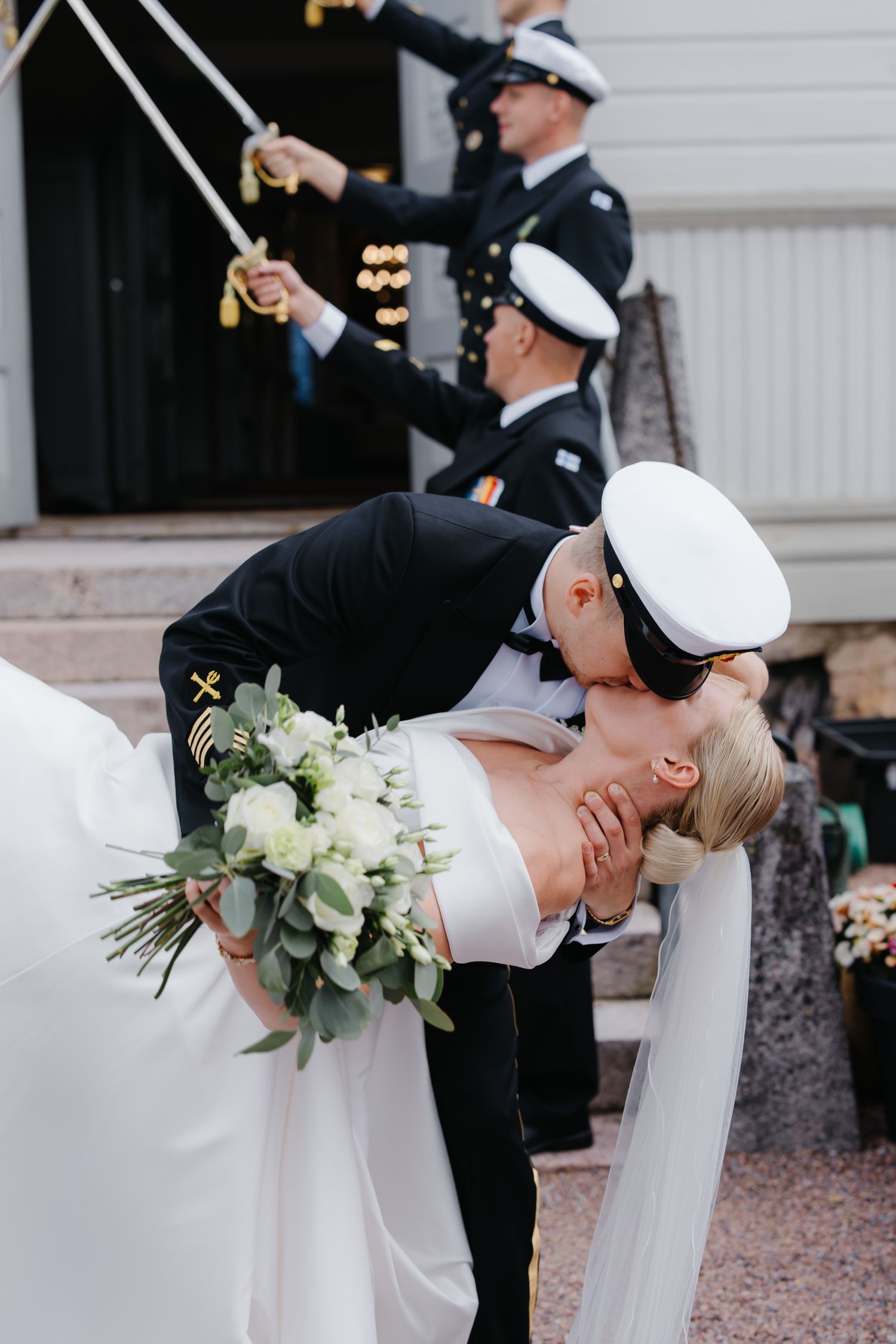 A vintage black car serves as a backdrop for wedding photos in a sunlit outdoor setting with lush greenery.