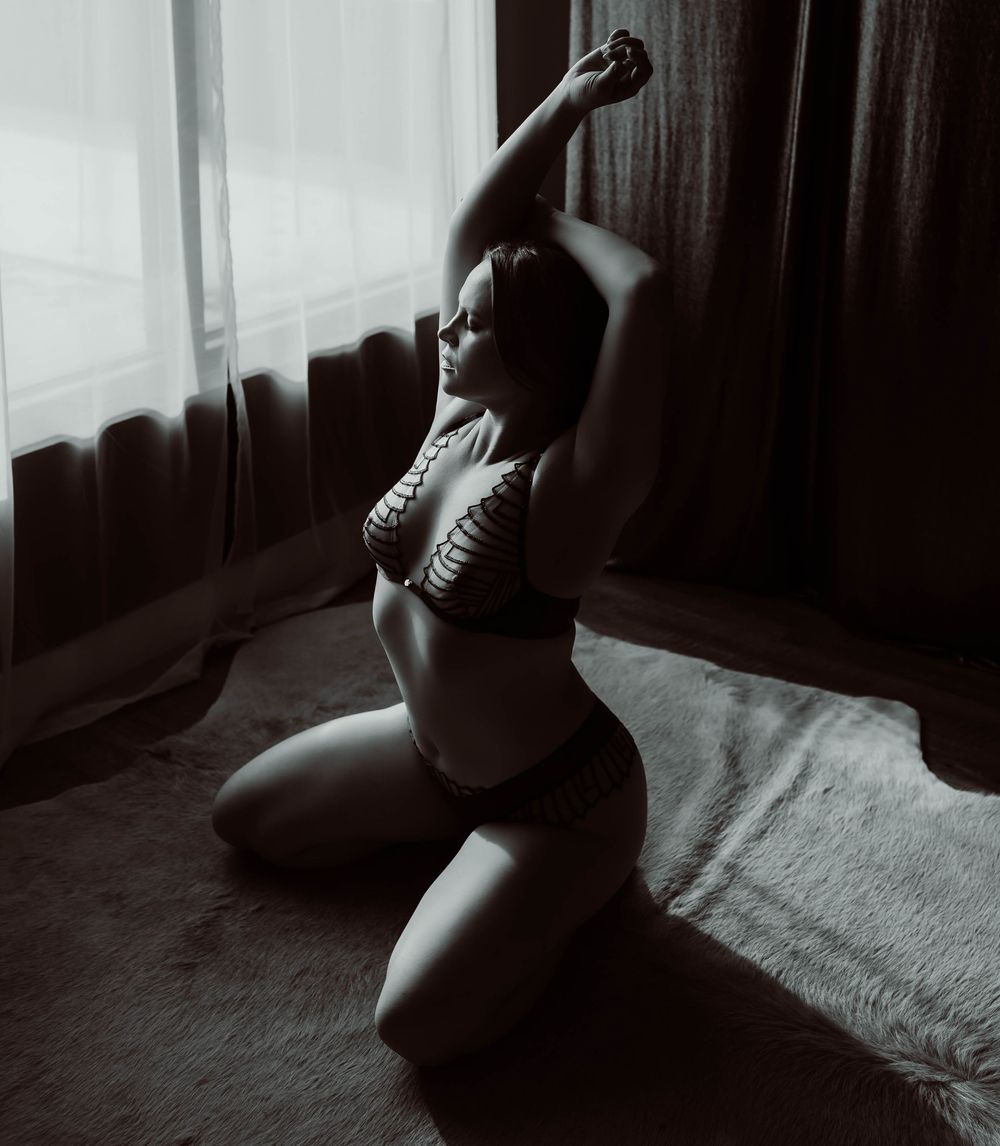 Black and white boudoir photo showing a woman in shadow sitting near a window.