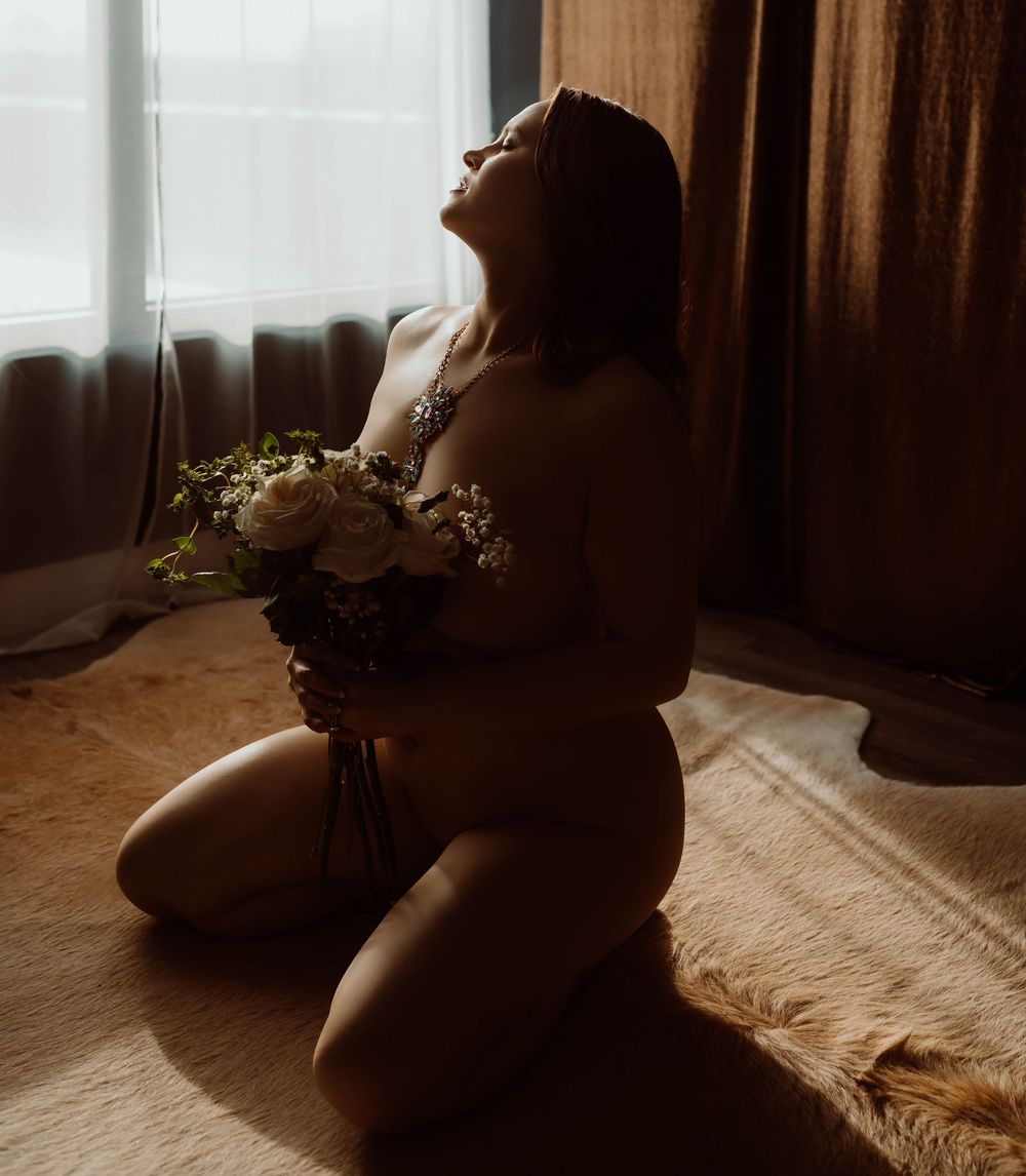Artistic boudoir silhouette of a figure kneeling with a bouquet of flowers by a sunlit window with Boudoir by KJ.