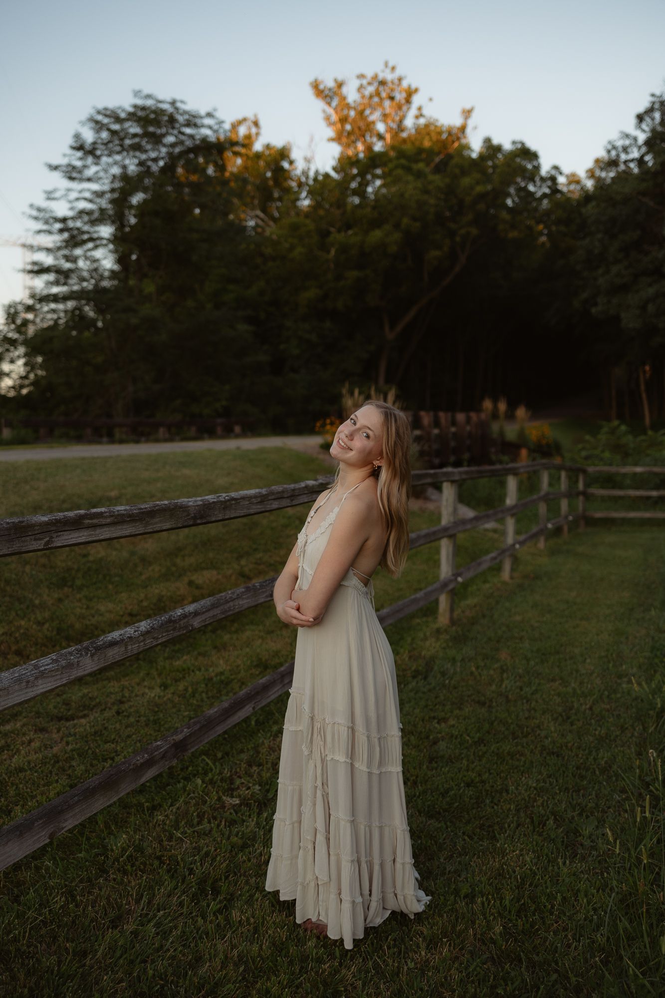A person in a flowy cream dress poses gracefully by a rustic wooden fence at sunset in a grassy field.