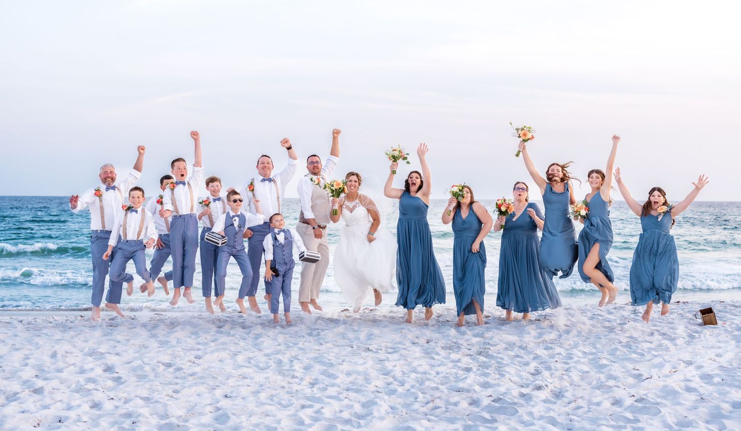 Wedding party wearing white and navy blue outfits poses together on a sunny beach during sunset celebration.