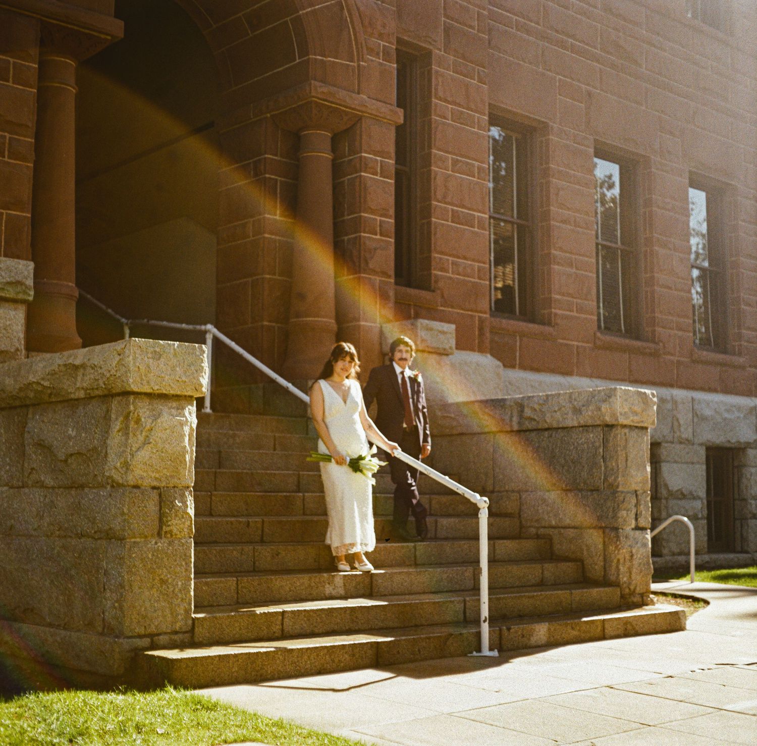 Bride and groom descend stone steps of a historic brick building bathed in golden sunlight on their wedding day.