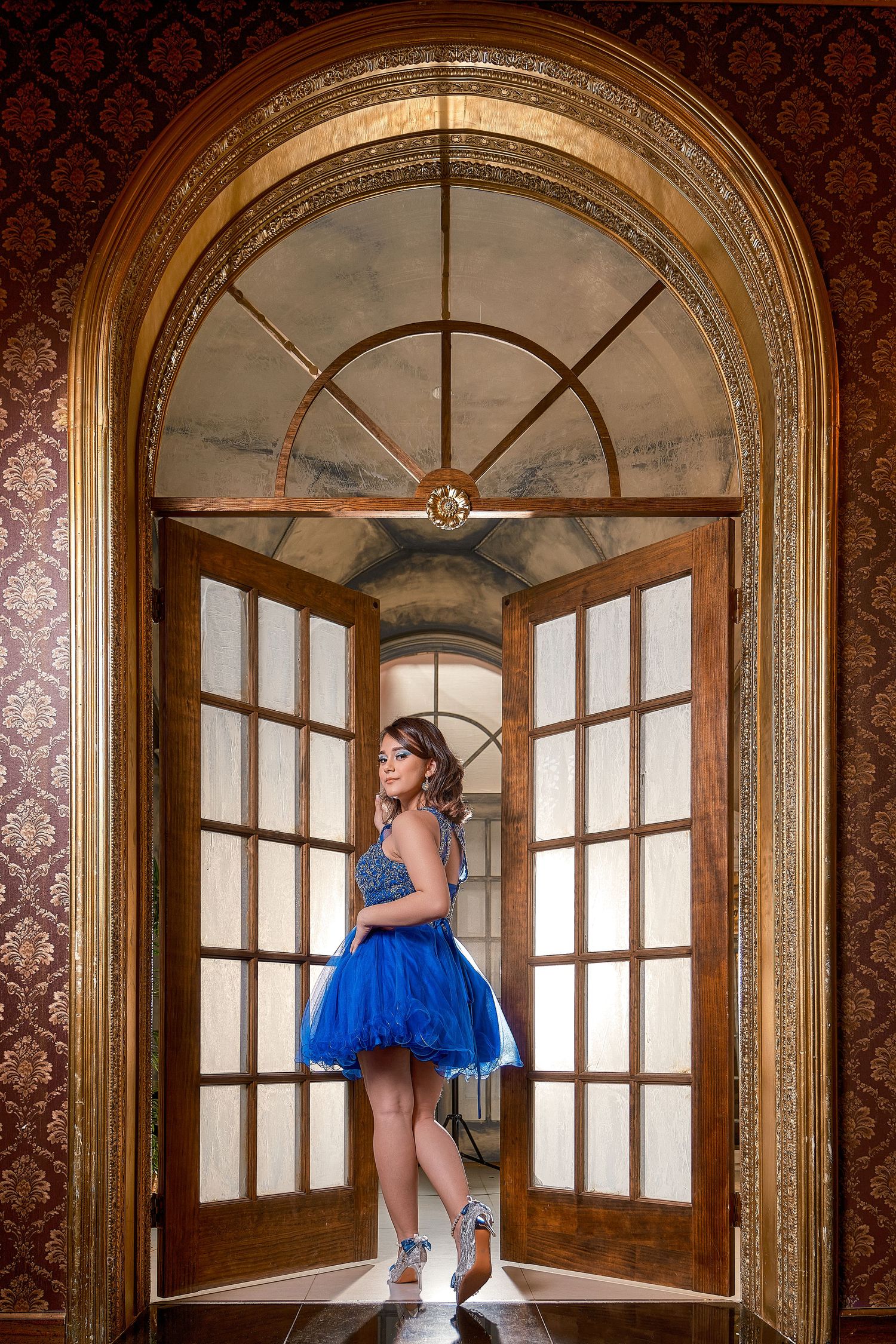 A figure in a blue tulle dress poses gracefully in an elegant room with ornate arched windows and damask wallpaper.