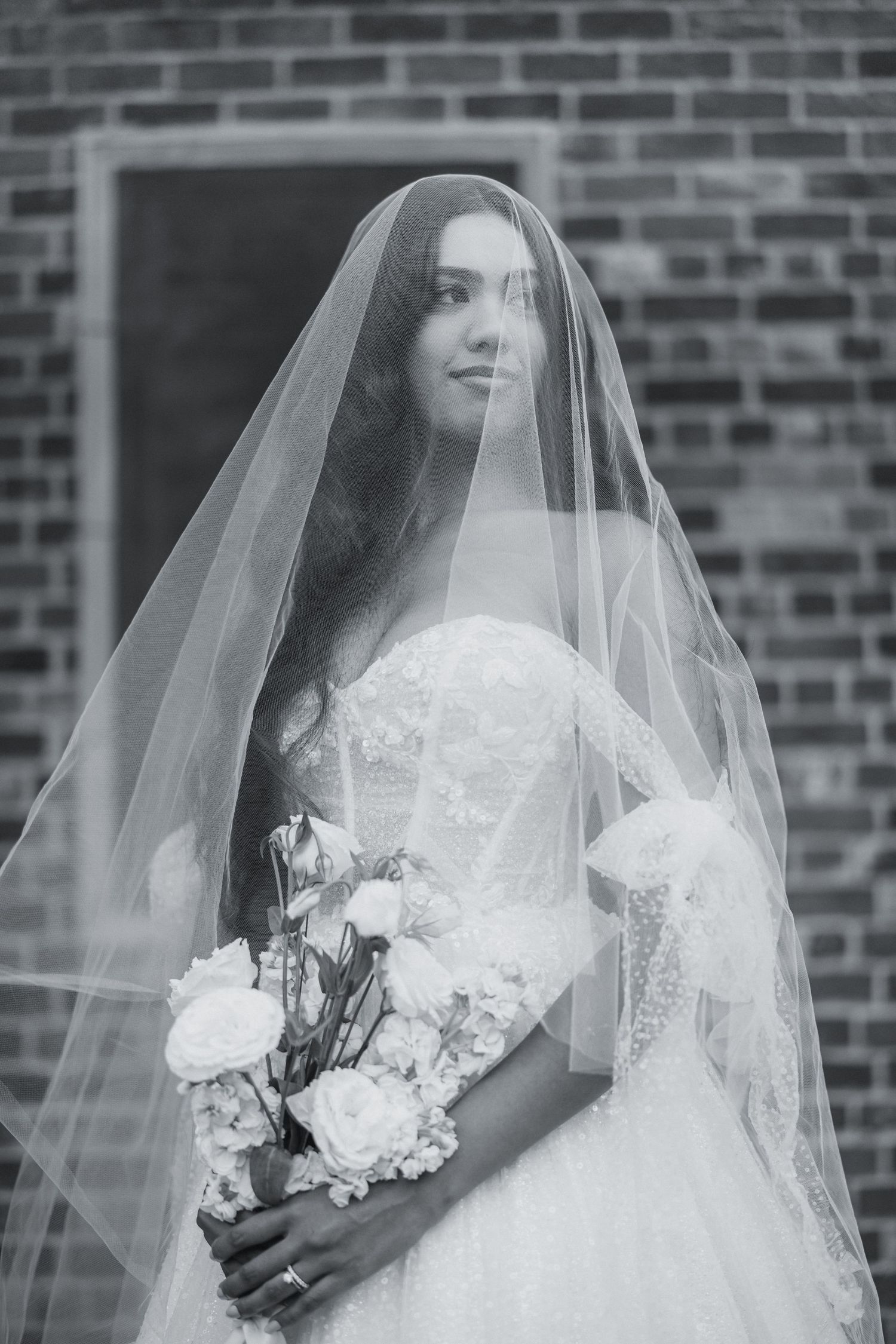 Artistic black and white bridal portraits with delicate veil and bouquet against brick wall.