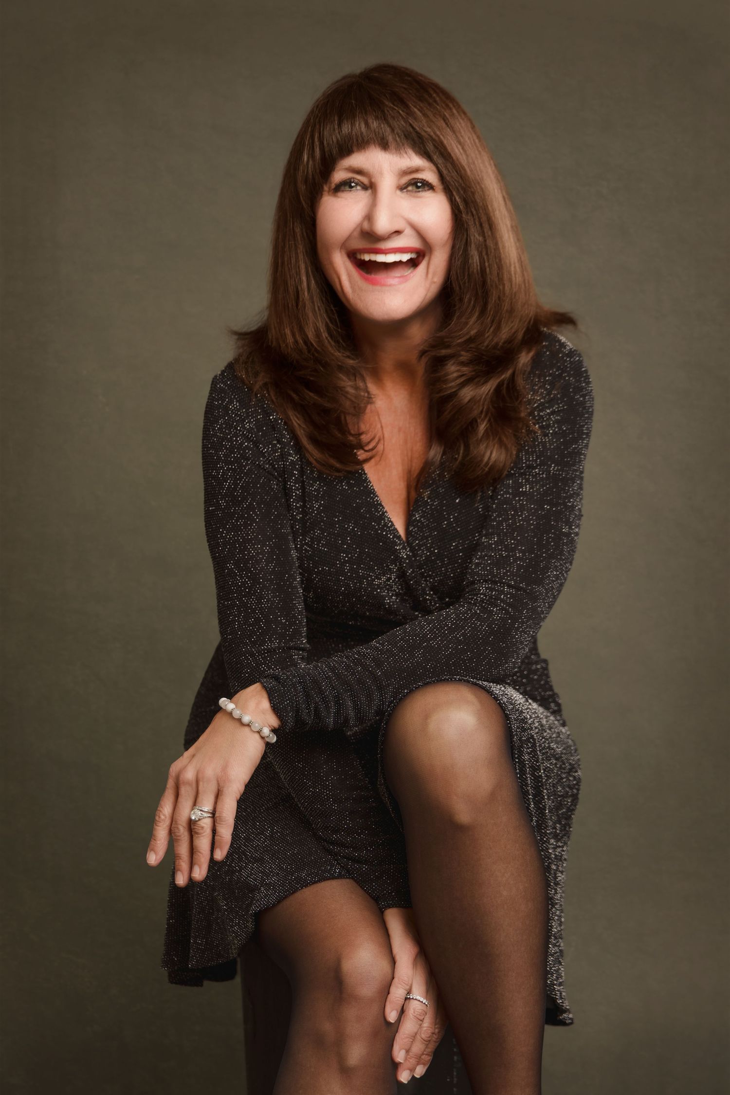 Smiling person in elegant black lace dress with jewelry sitting in casual pose against dark background.