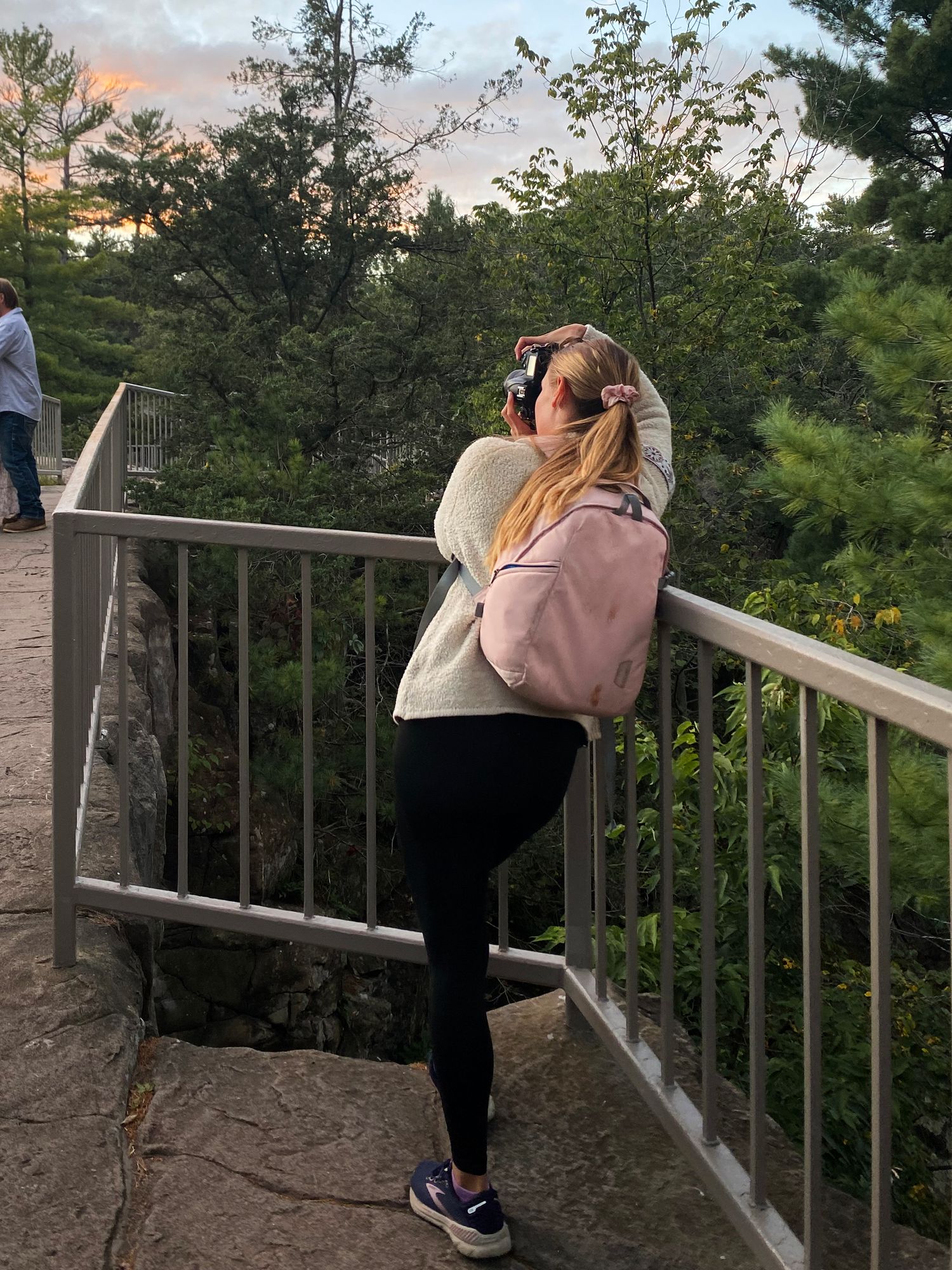 Someone wearing a pink sweater takes a photo from a scenic overlook with metal railings at sunset among pine trees.