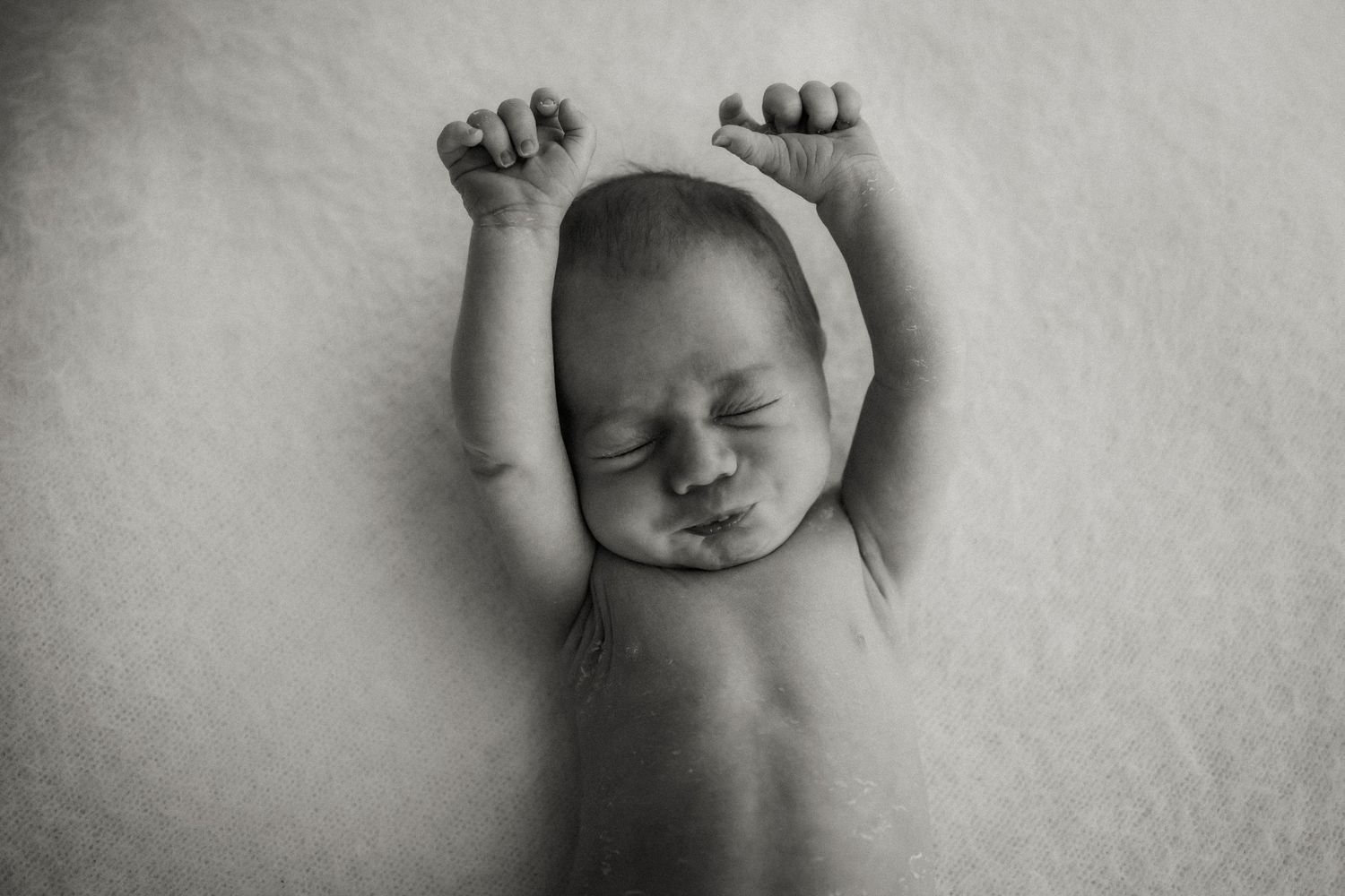 Baby in white onesie lying on white bedding viewed from above.