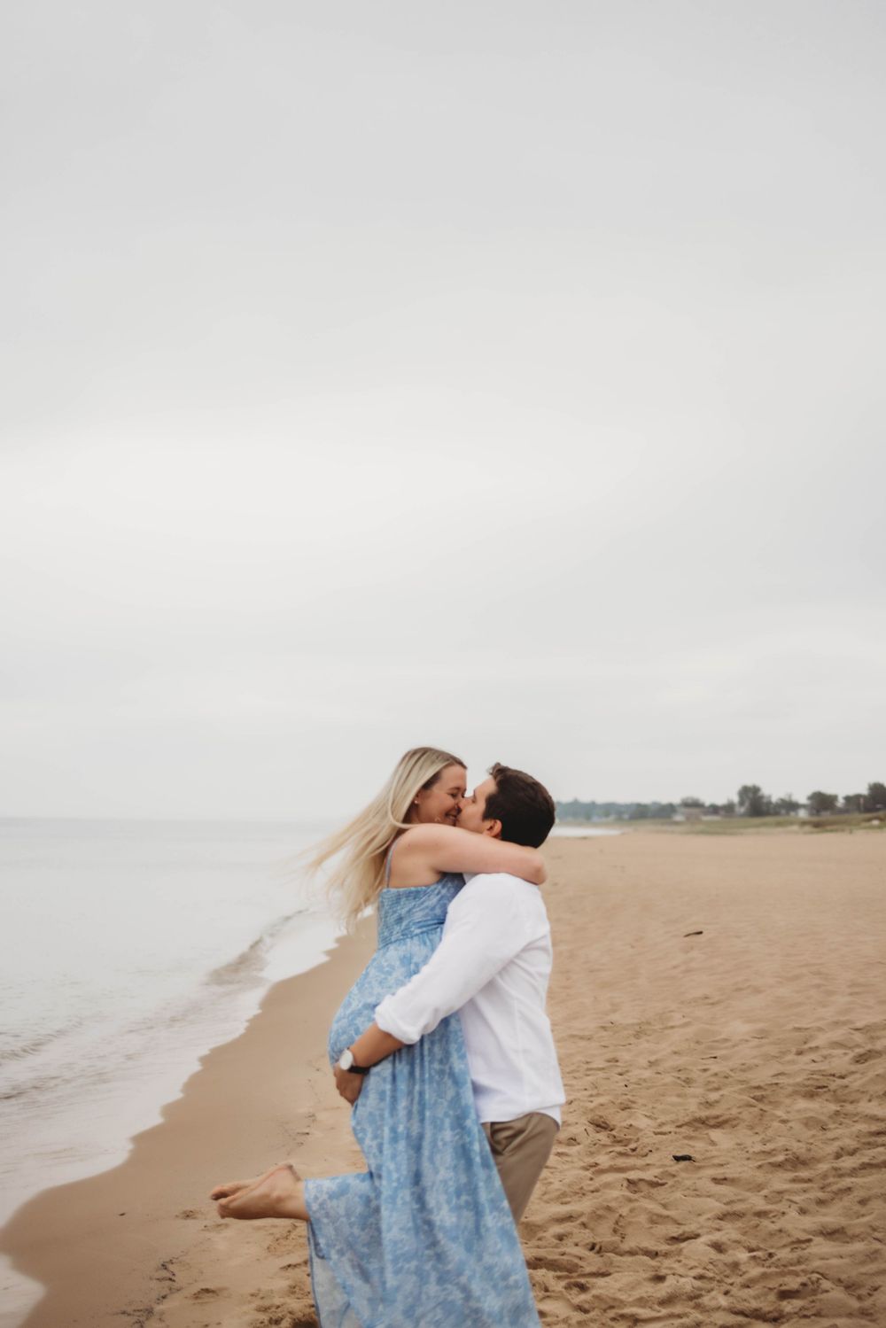 Romantic couple embracing and kissing on a sandy beach with overcast sky and water in the background.