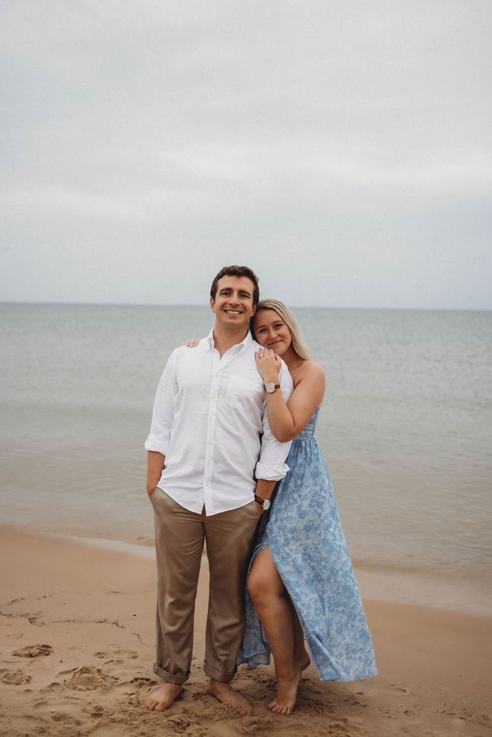 A couple shares an intimate moment on a sandy beach during an overcast day with waves in the background.