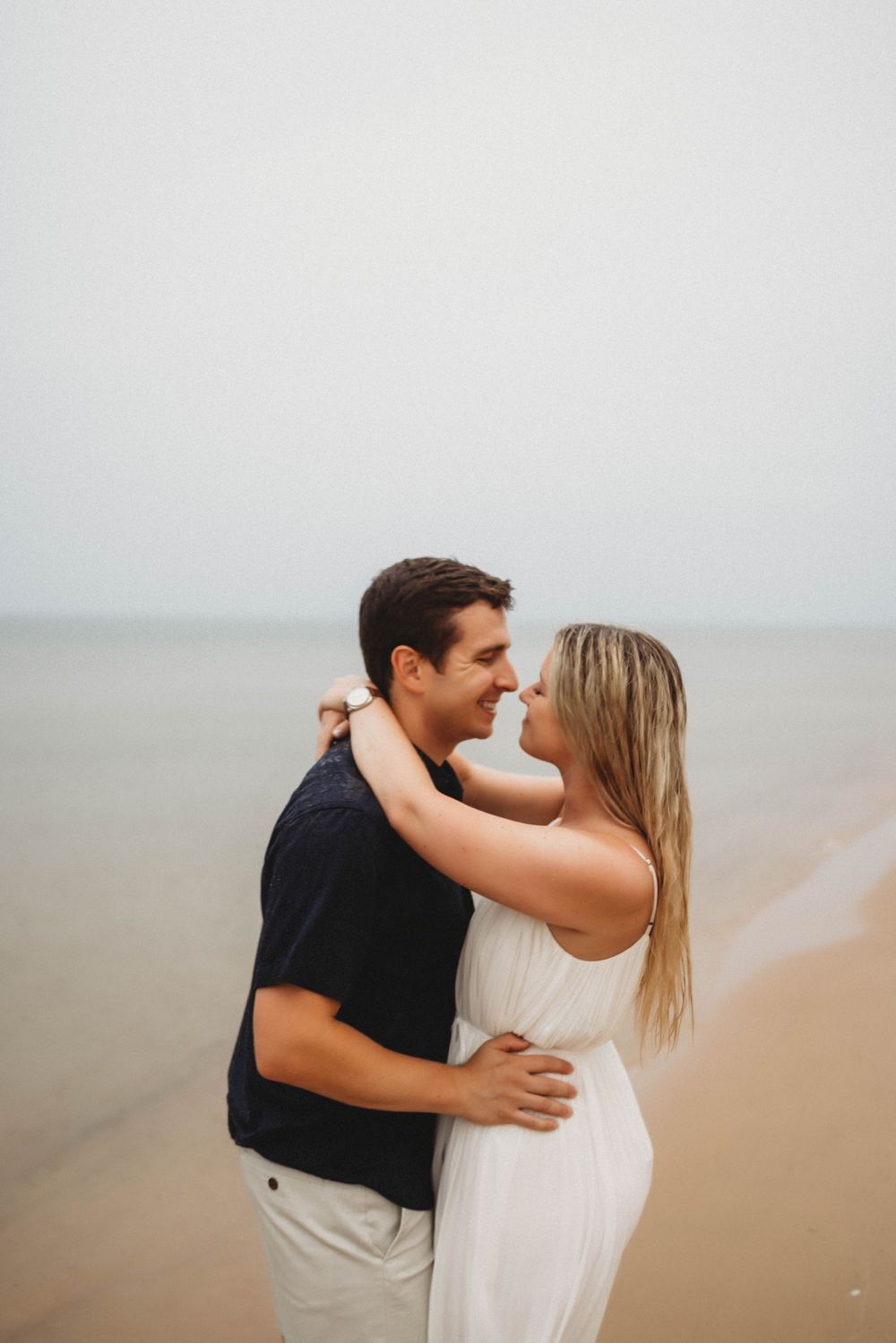 A couple share intimate embraces and kisses while walking along a misty beach at sunset.