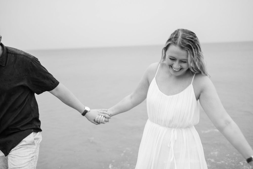 Black and white photo of a couple holding hands while walking along a misty beach coastline.