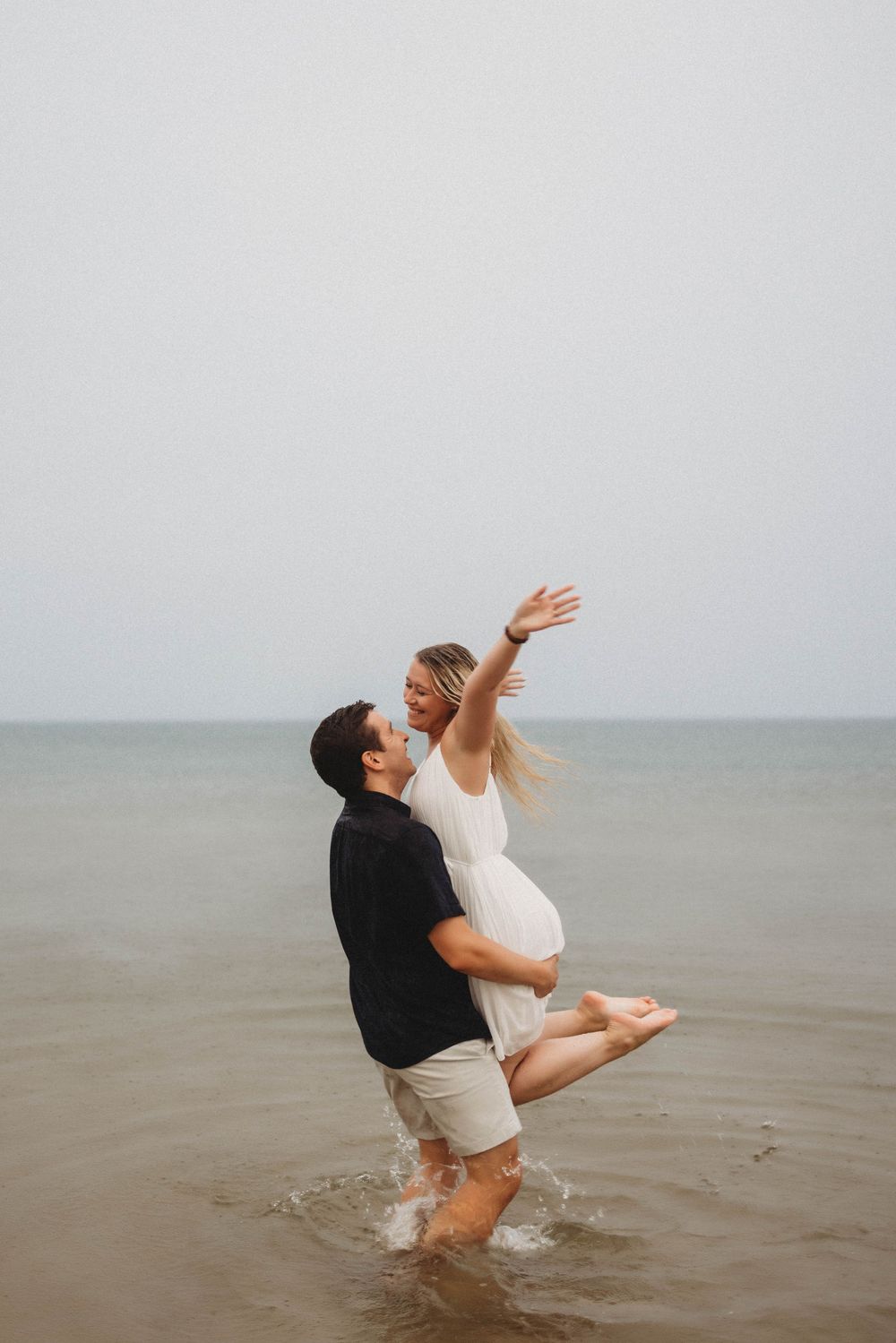Couple embraces joyfully in shallow ocean water during romantic beach photoshoot at dusk.