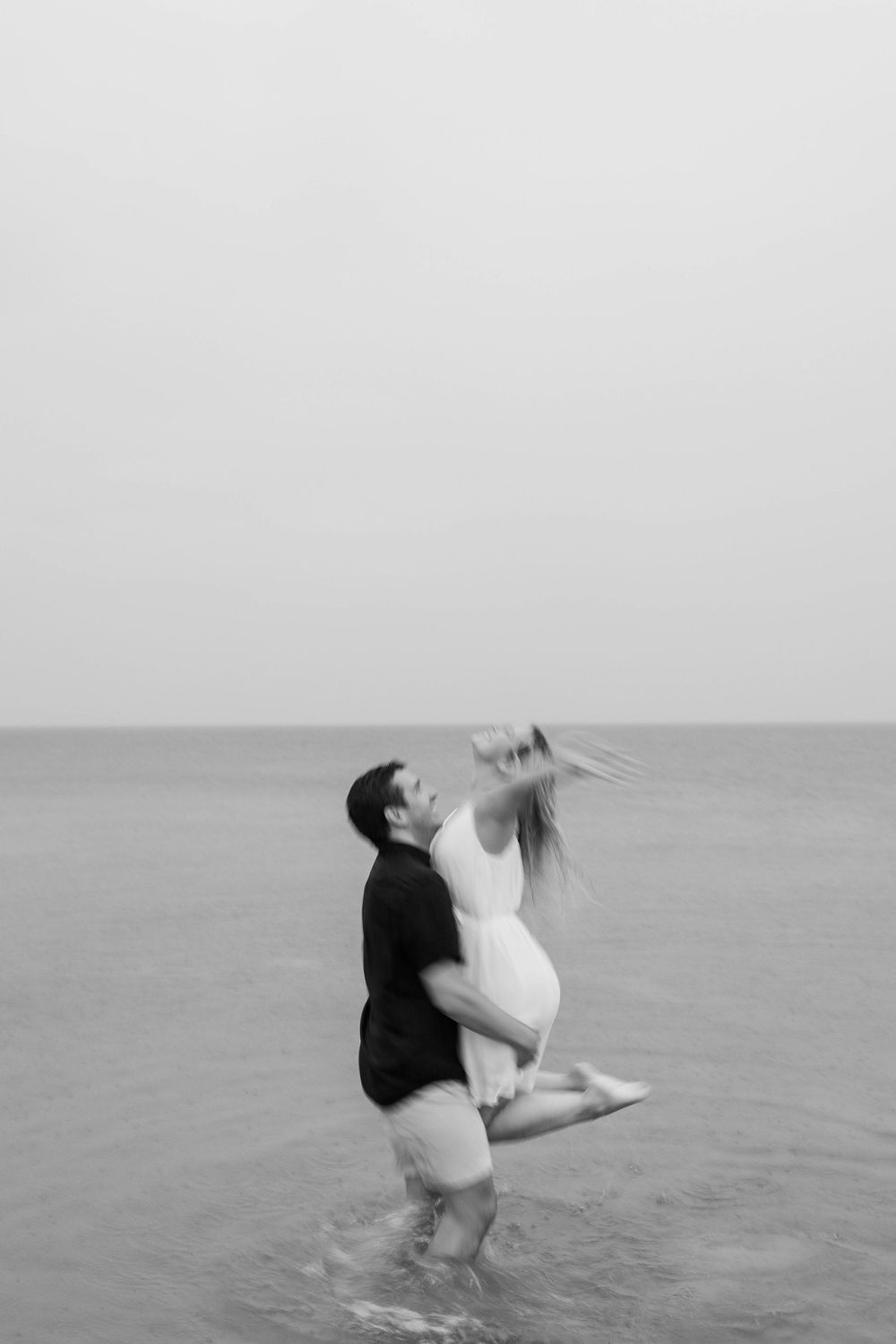 A couple dances joyfully in shallow ocean water, creating a romantic black and white moment at the beach.