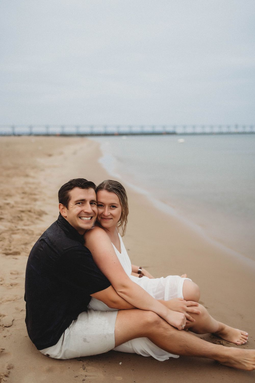 A romantic couple lounges together on a sandy beach with a pier visible in the distant background.