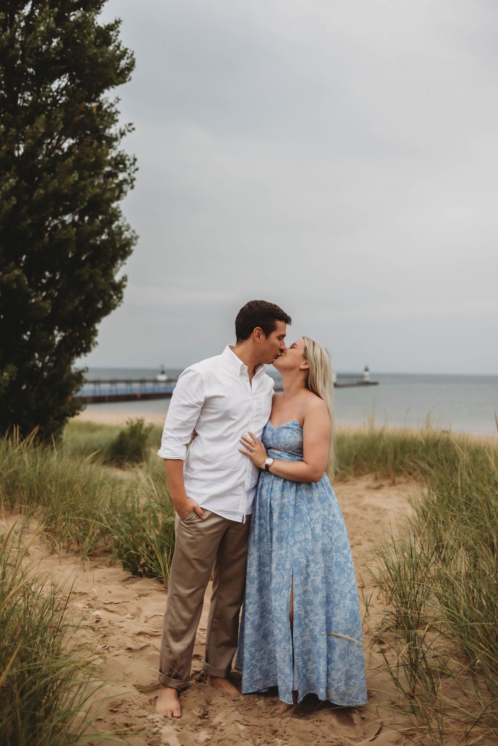 A couple shares an intimate moment among coastal sand dunes with the ocean visible in the background.