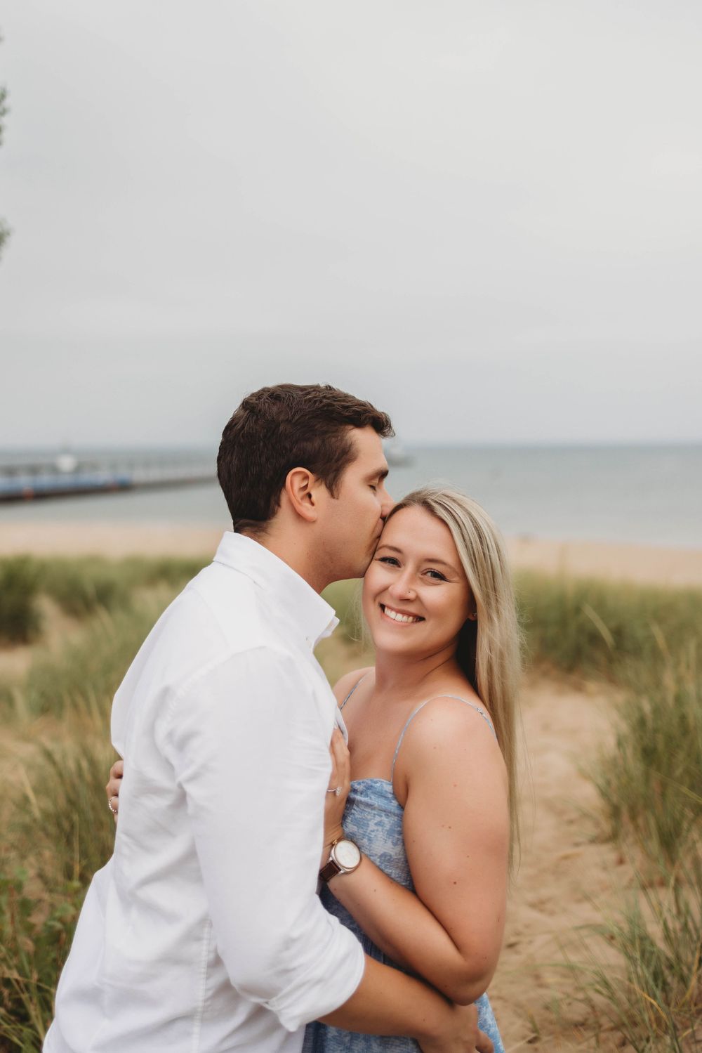 A romantic couple embraces on a beach with dunes and ocean in the background.