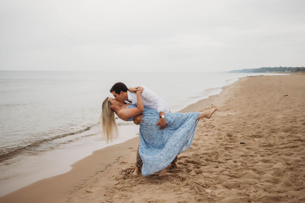 Romantic couple dips and dances together on a misty beach shoreline with woman wearing flowing blue dress.