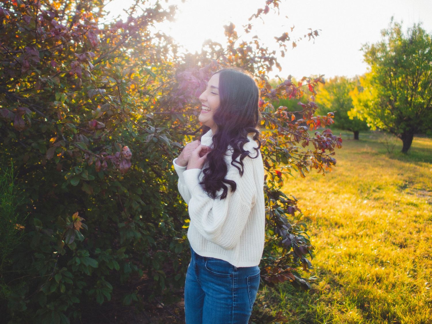 A young woman in a white sweater and jeans stands in a sunlit garden with autumn foliage during golden hour.