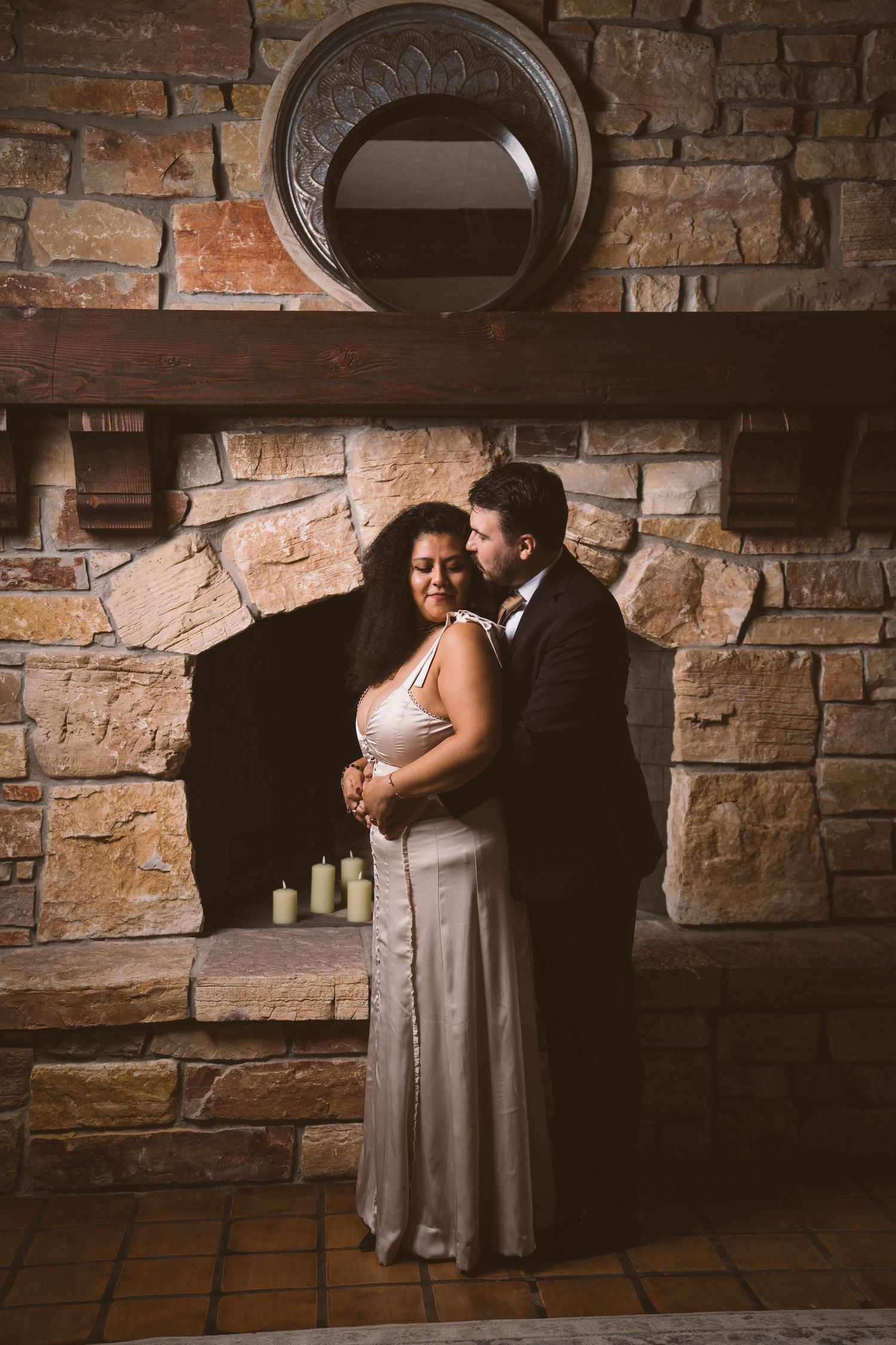 A romantic couple embraces against a rustic brick wall backdrop in a dimly lit setting.