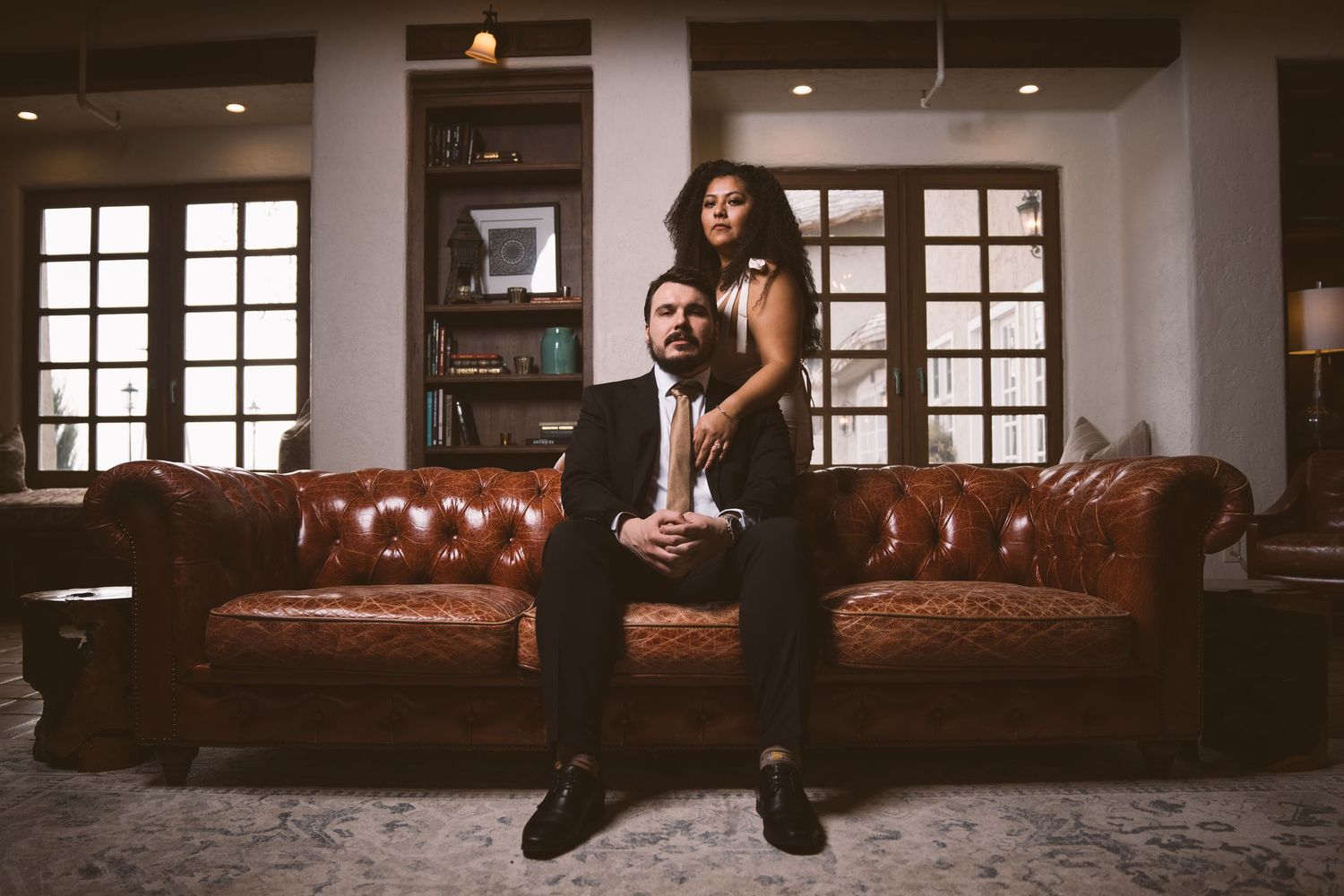 A couple lounges intimately on a brown leather couch in an elegant room with windows.