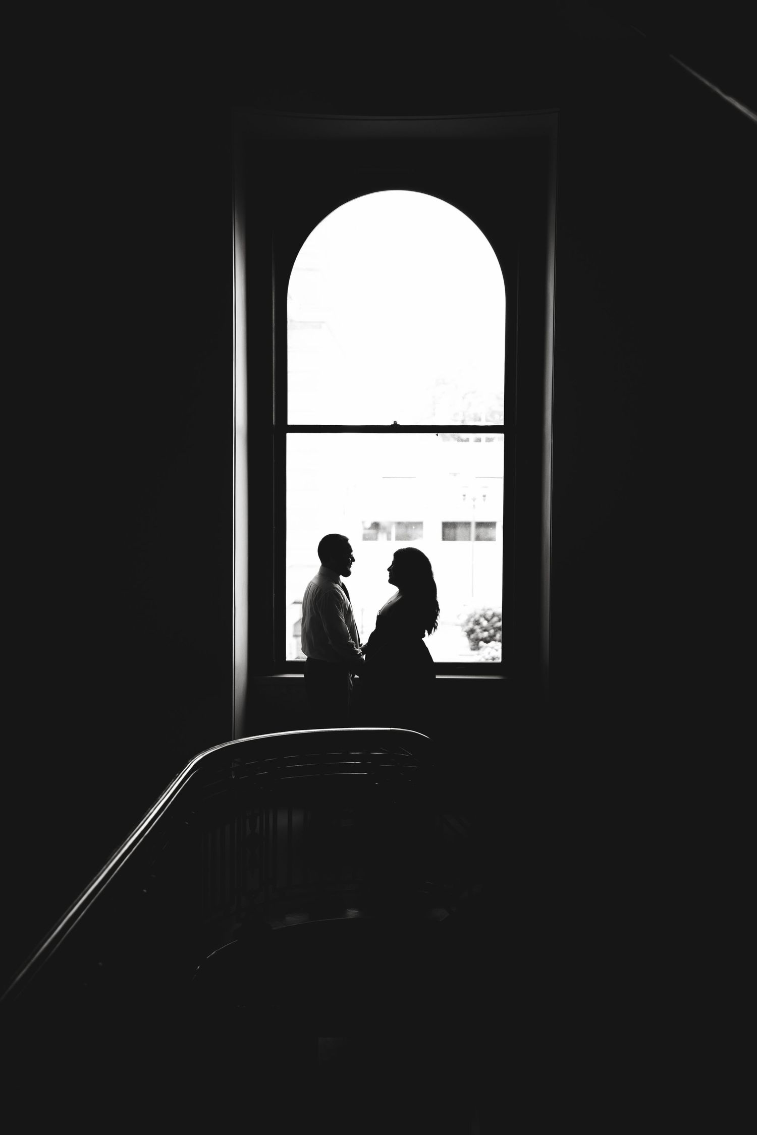 Silhouettes of two people standing by arched window in dark stairwell.