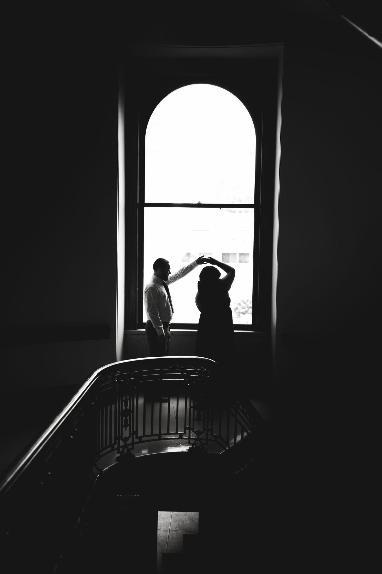 Two silhouettes dance by large arched window in dramatic black and white composition.