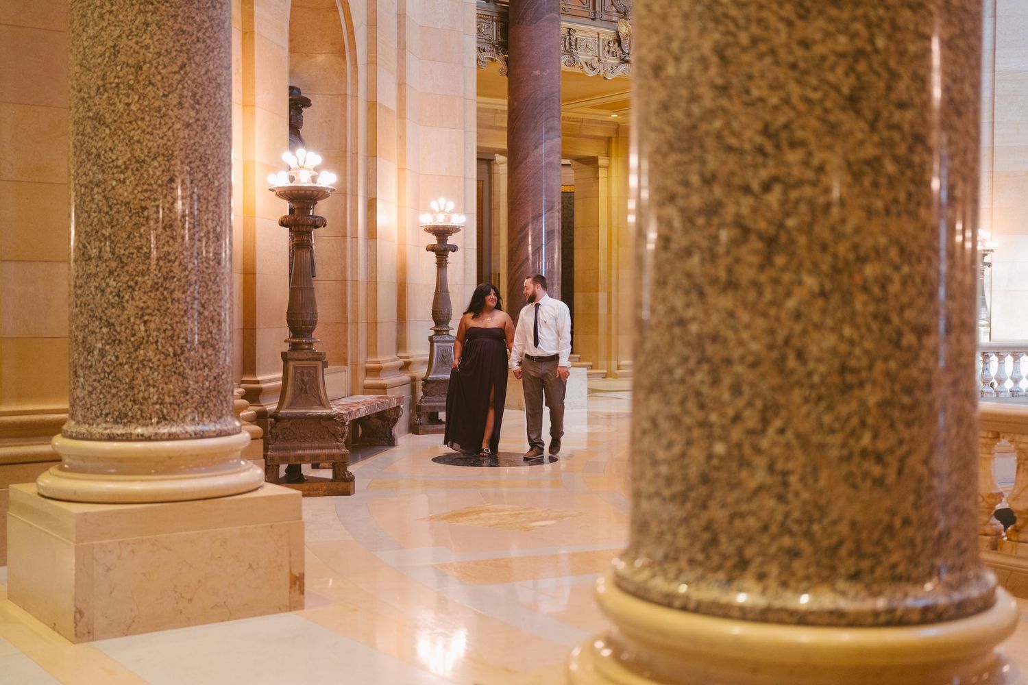 Couple walking through elegant hallway with marble columns and ornate lighting.