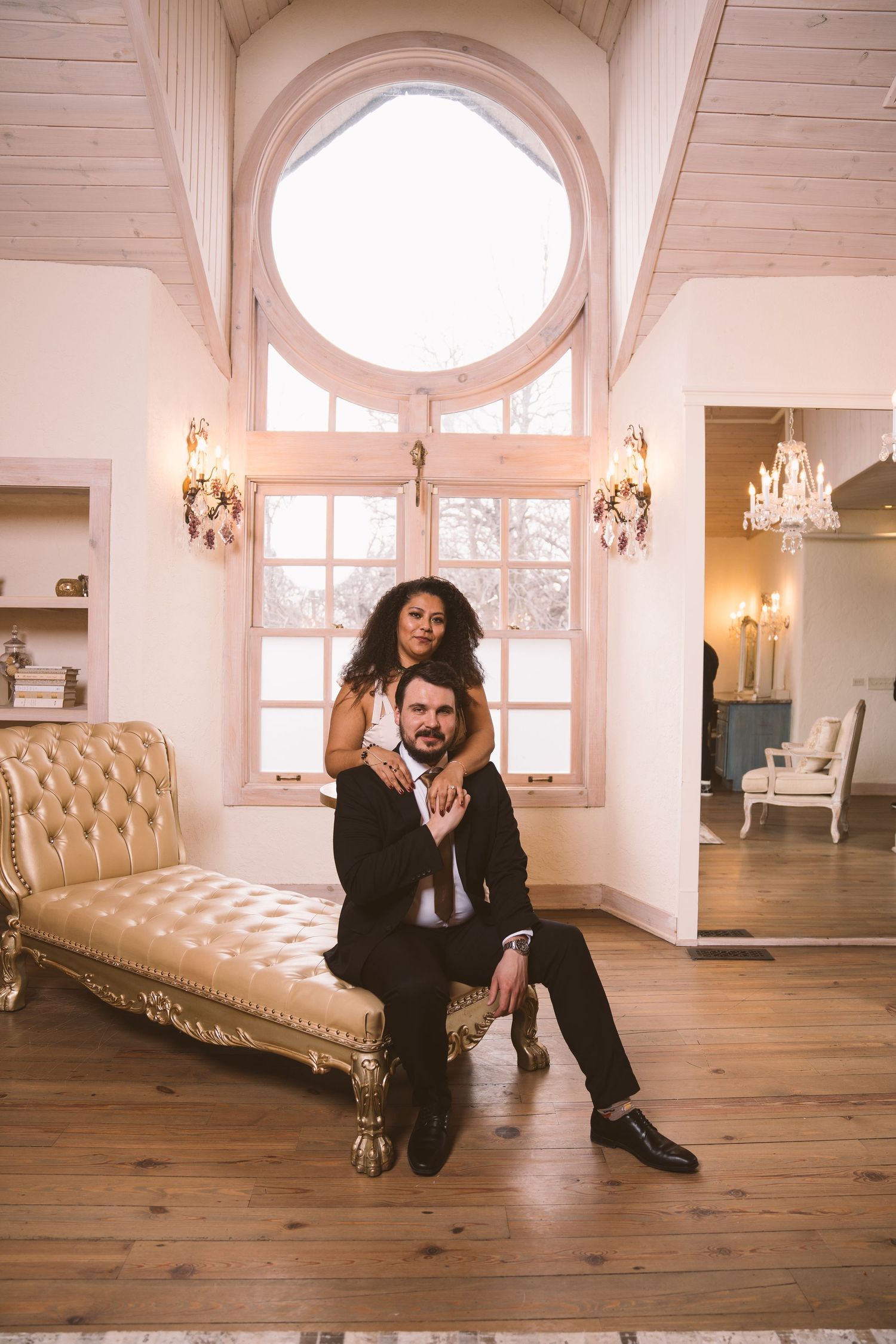 A couple poses on hardwood floors in an elegant white room with arched windows and chandeliers.