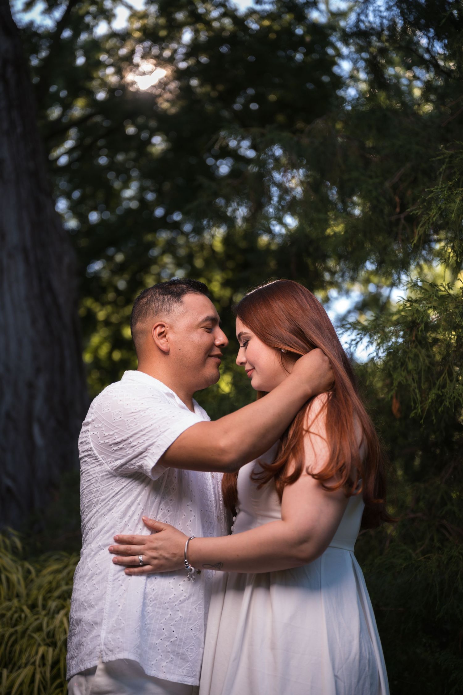 A romantic couple in white outfits shares tender moments in a wooded outdoor setting during a photo session.