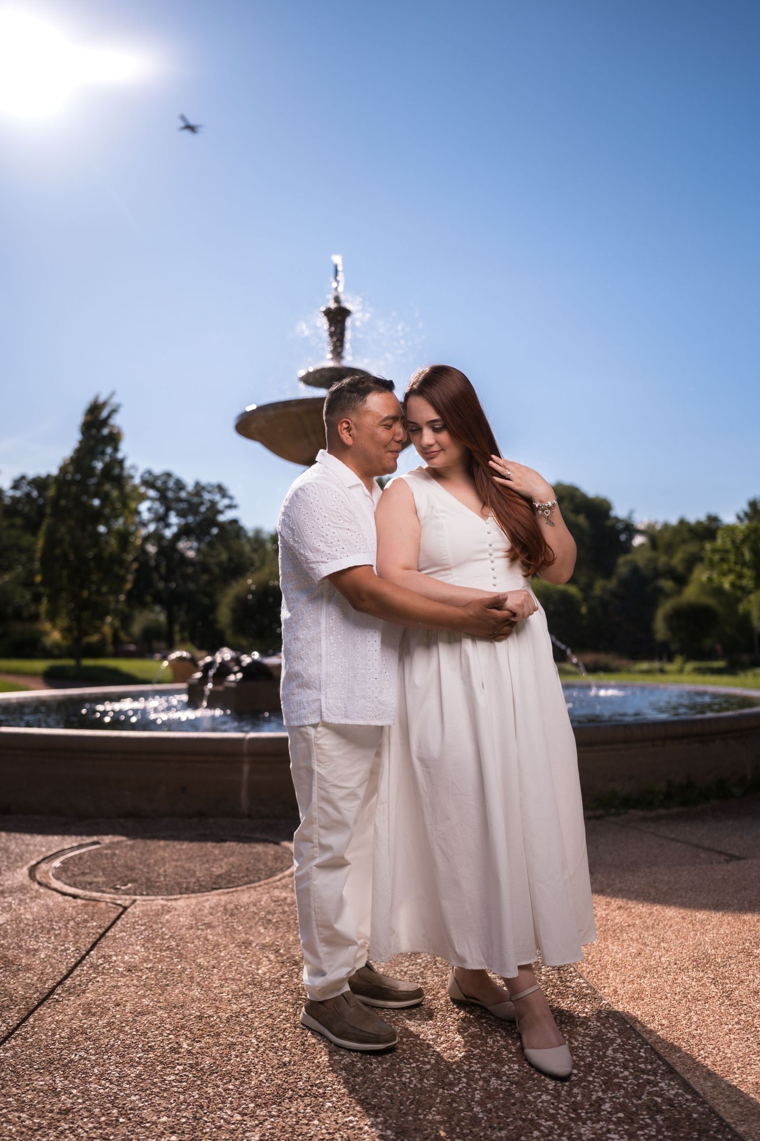 A couple dressed in white embraces on a paved garden path on a sunny afternoon.