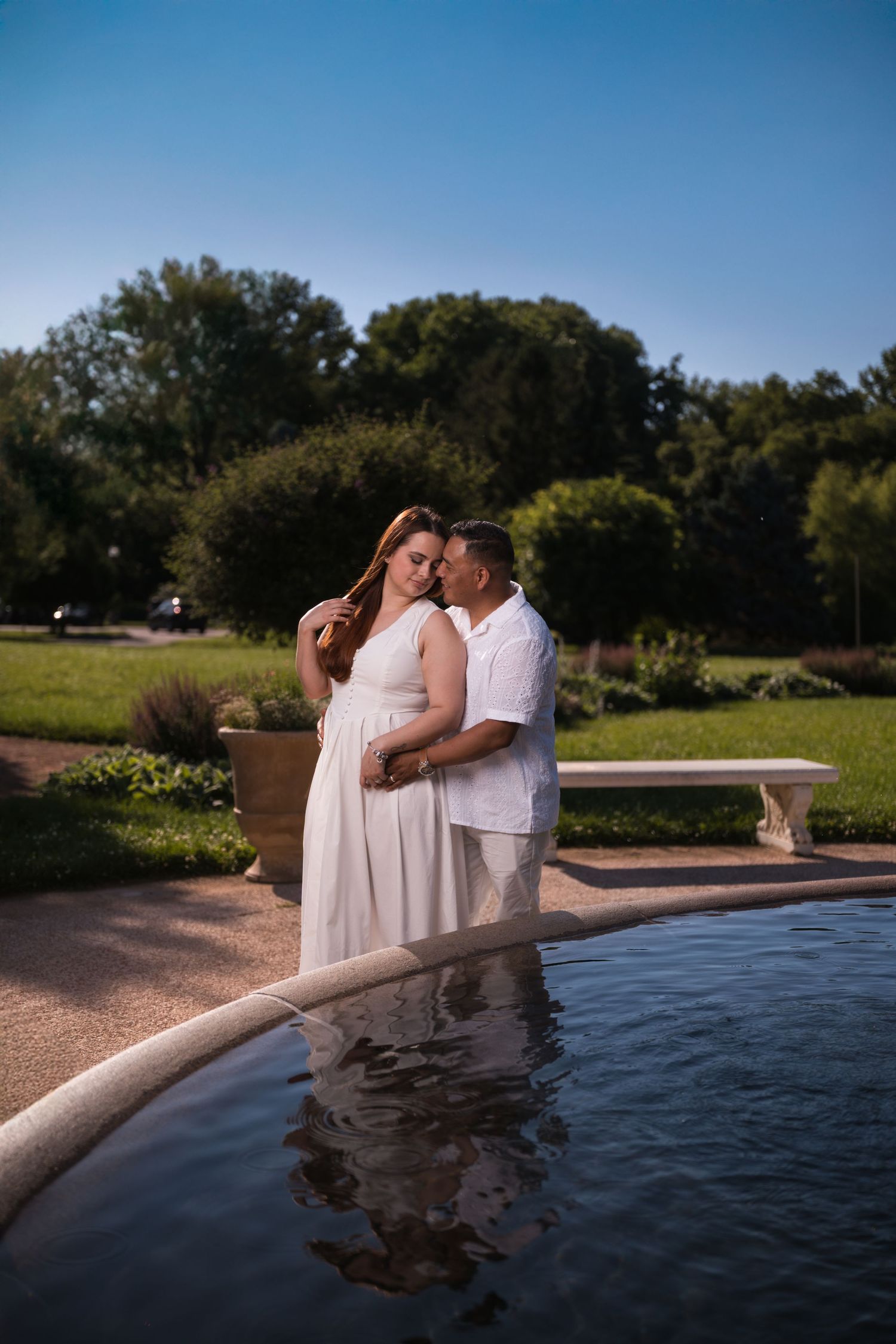 A couple in white clothing poses romantically by a reflective pool in a garden setting.