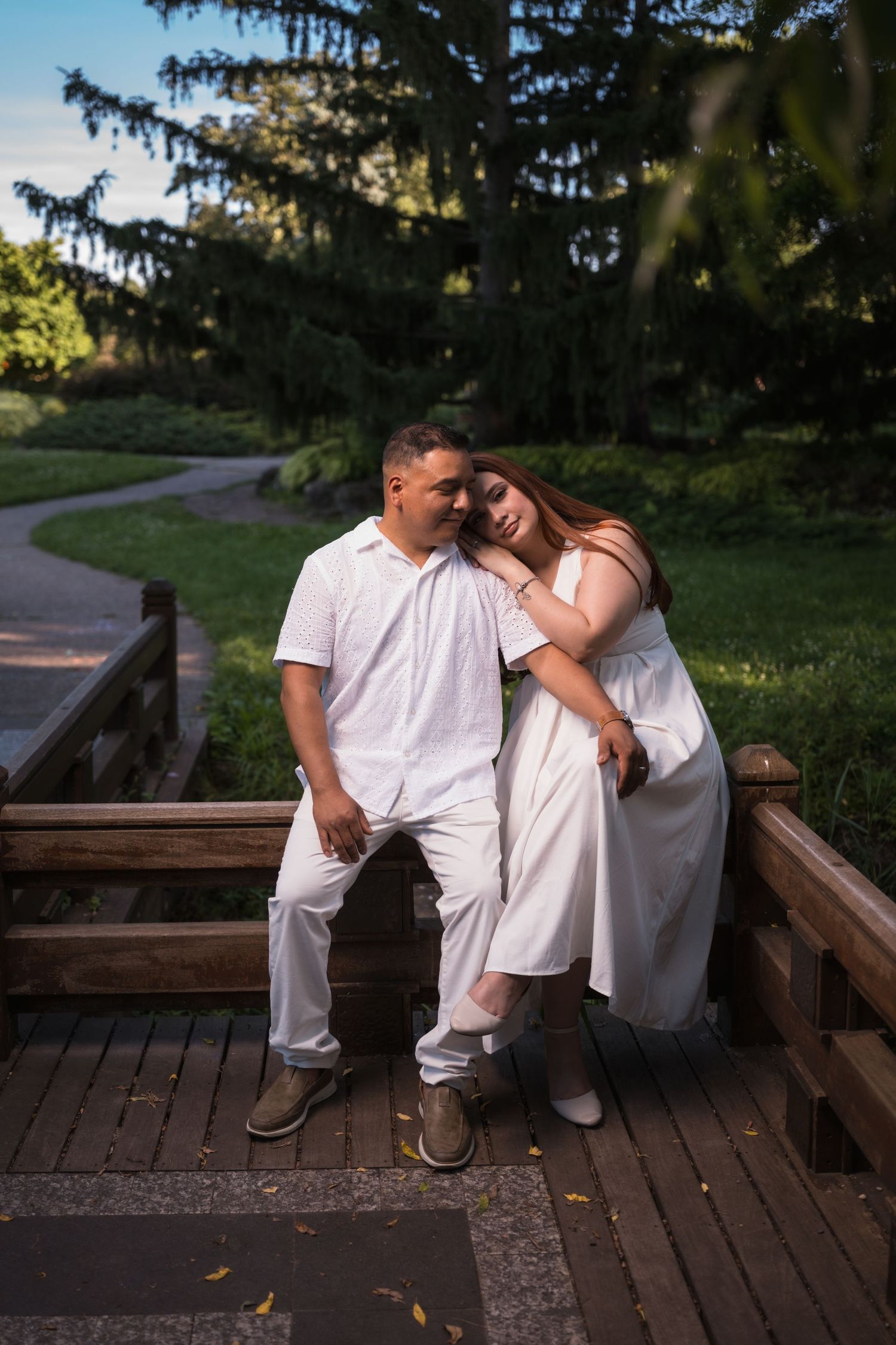 A couple in white outfits shares tender moments on a wooden deck surrounded by evergreen trees.