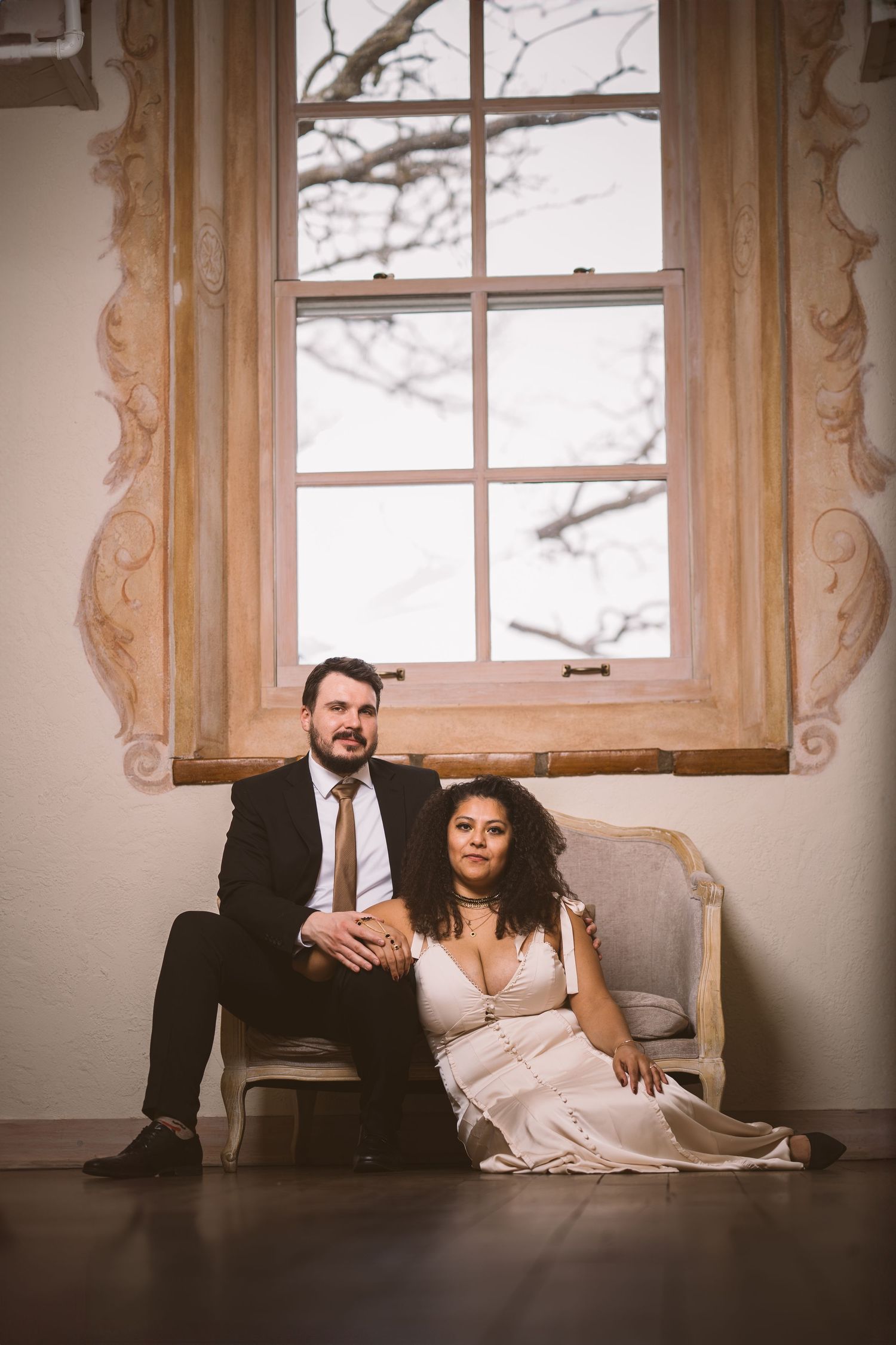 A couple relaxes by large vintage windows in a bright, airy room.