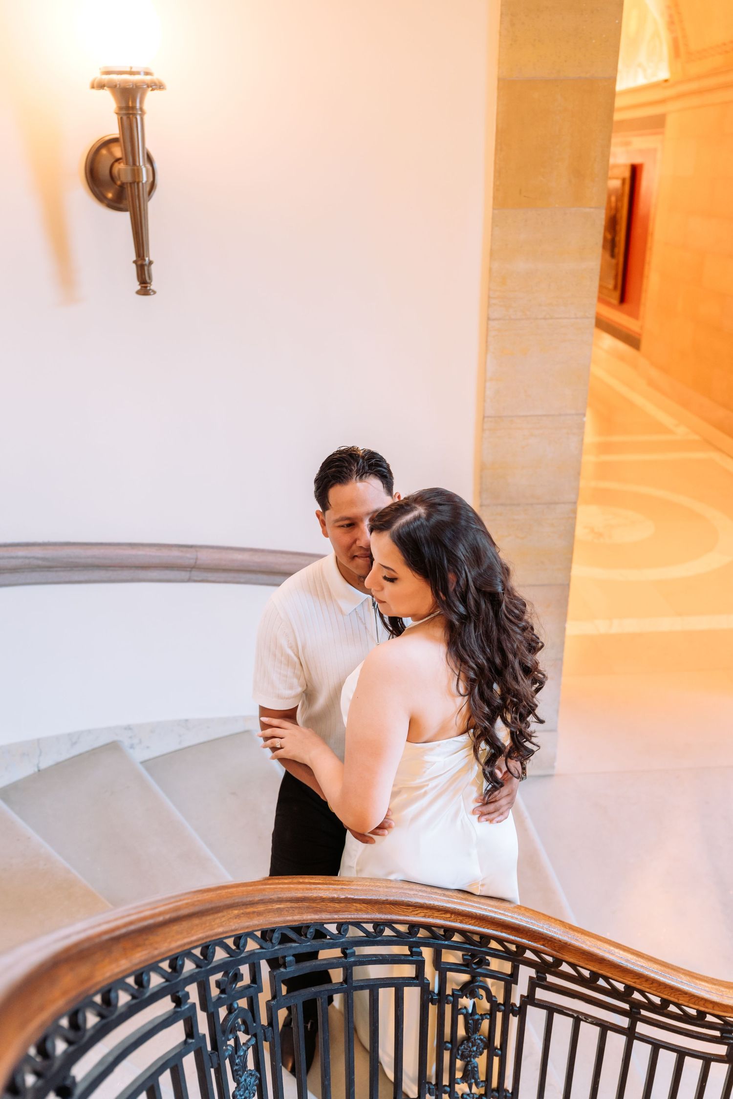Couple embraces on an elegant curved staircase with ornate railings in warm lighting.