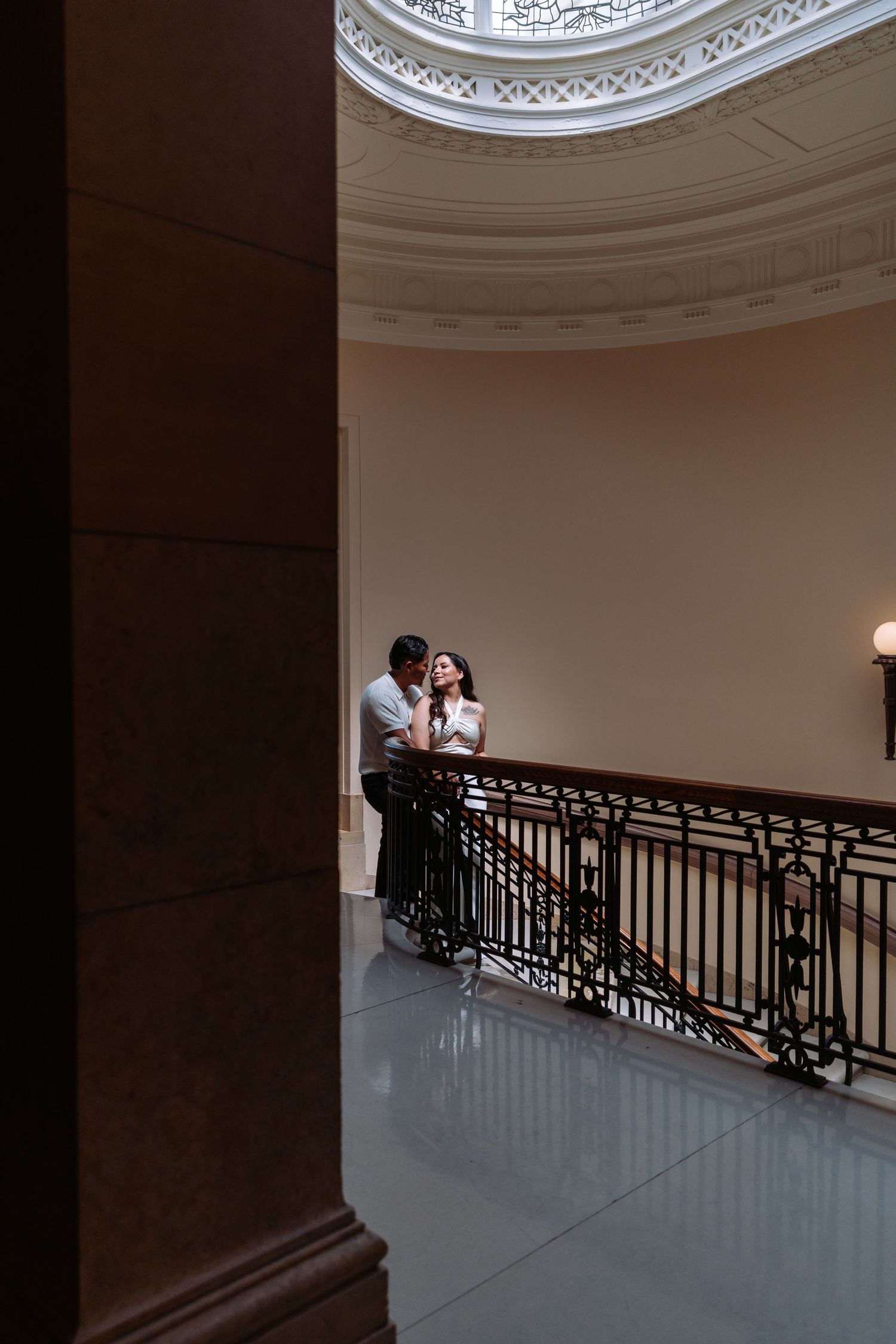 Grand curved staircase with ornate iron railings provides dramatic setting for couple's portrait.
