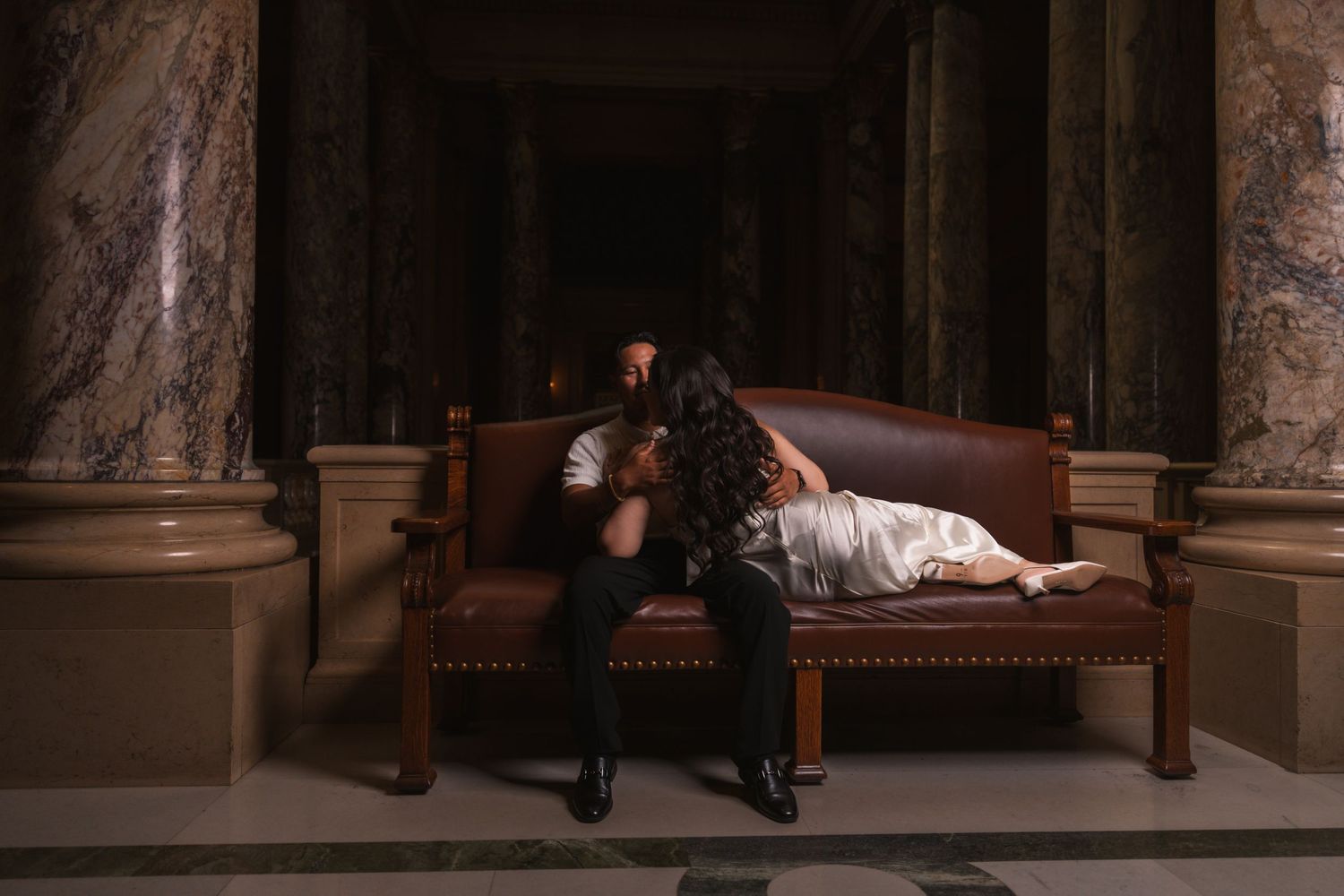 Couple poses dramatically on vintage bench in historic building hallway.