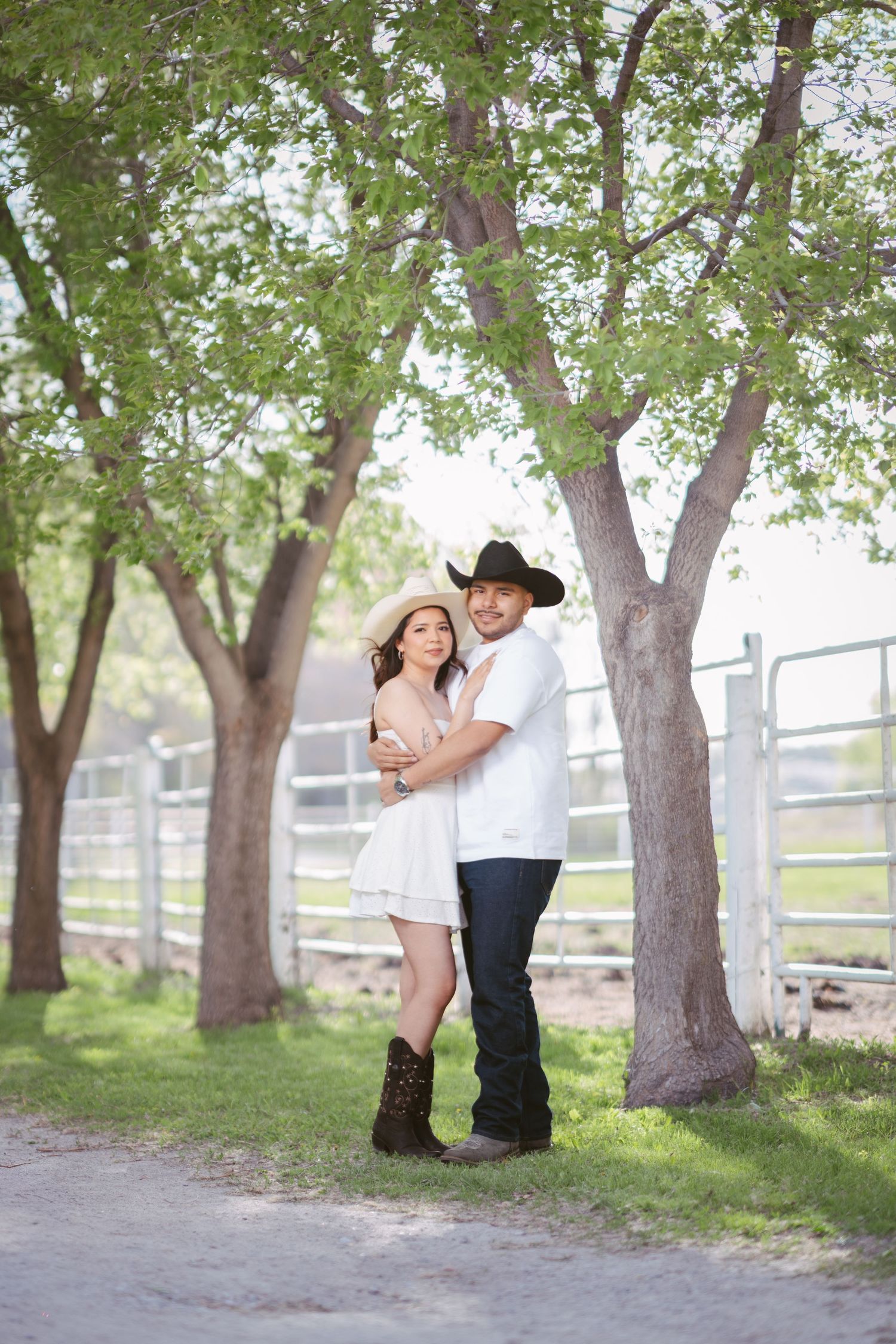 Couple in cowboy hats embracing under green trees on a ranch, woman in white dress and boots, man in jeans.
