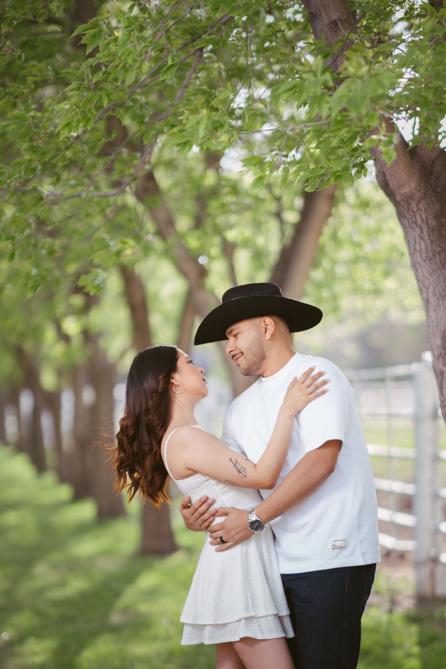 Couple embracing among green trees, woman in white dress looks up at man wearing black cowboy hat outdoors.