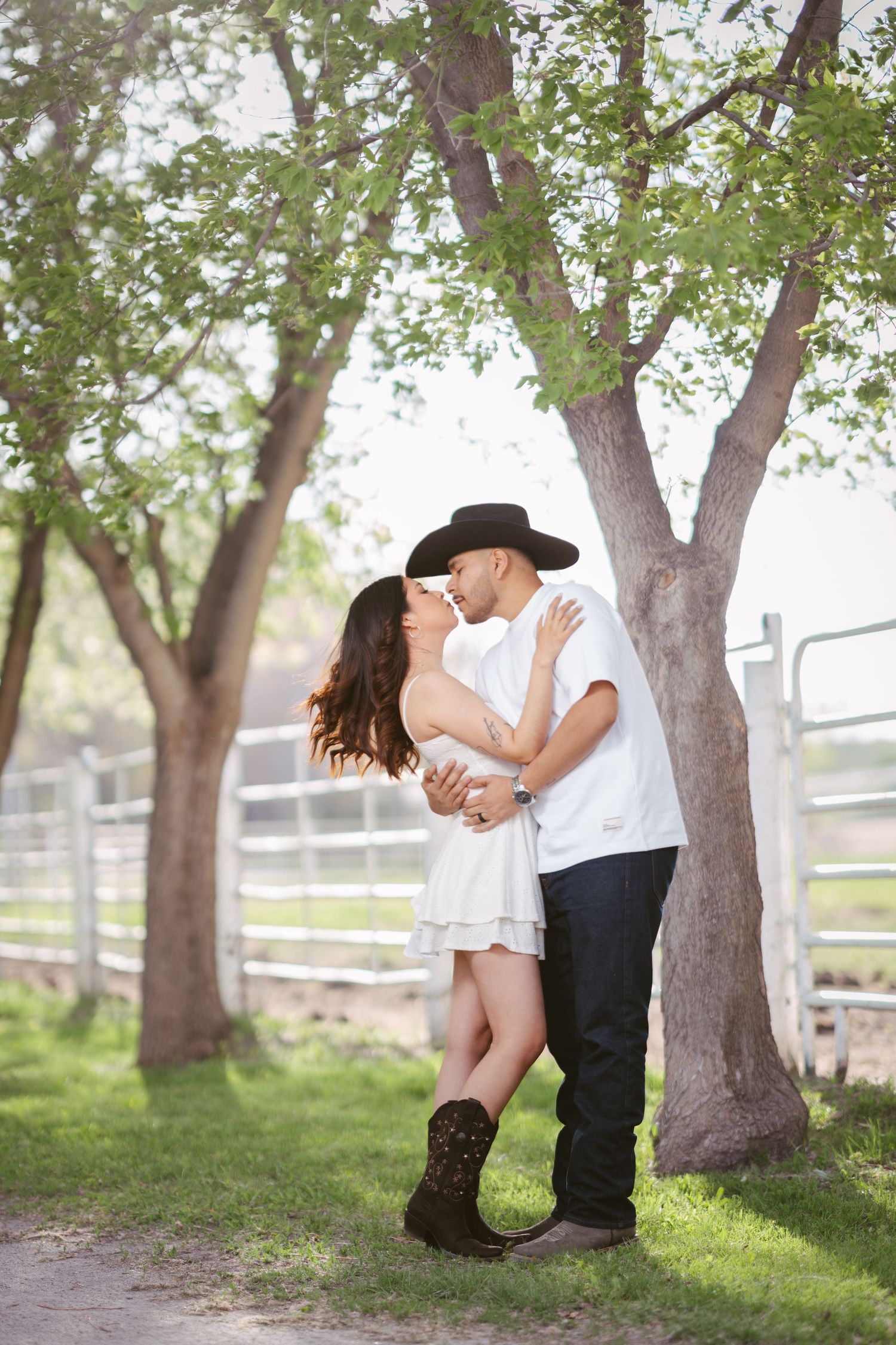 Couple sharing a romantic kiss under green trees, woman in white dress and cowboy boots, man in black cowboy hat.