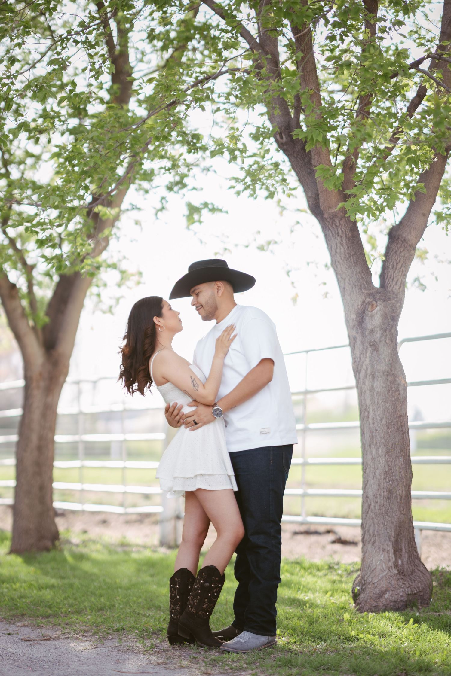 Couple in western wear embracing among trees, woman in white dress and cowboy boots, man in black hat.