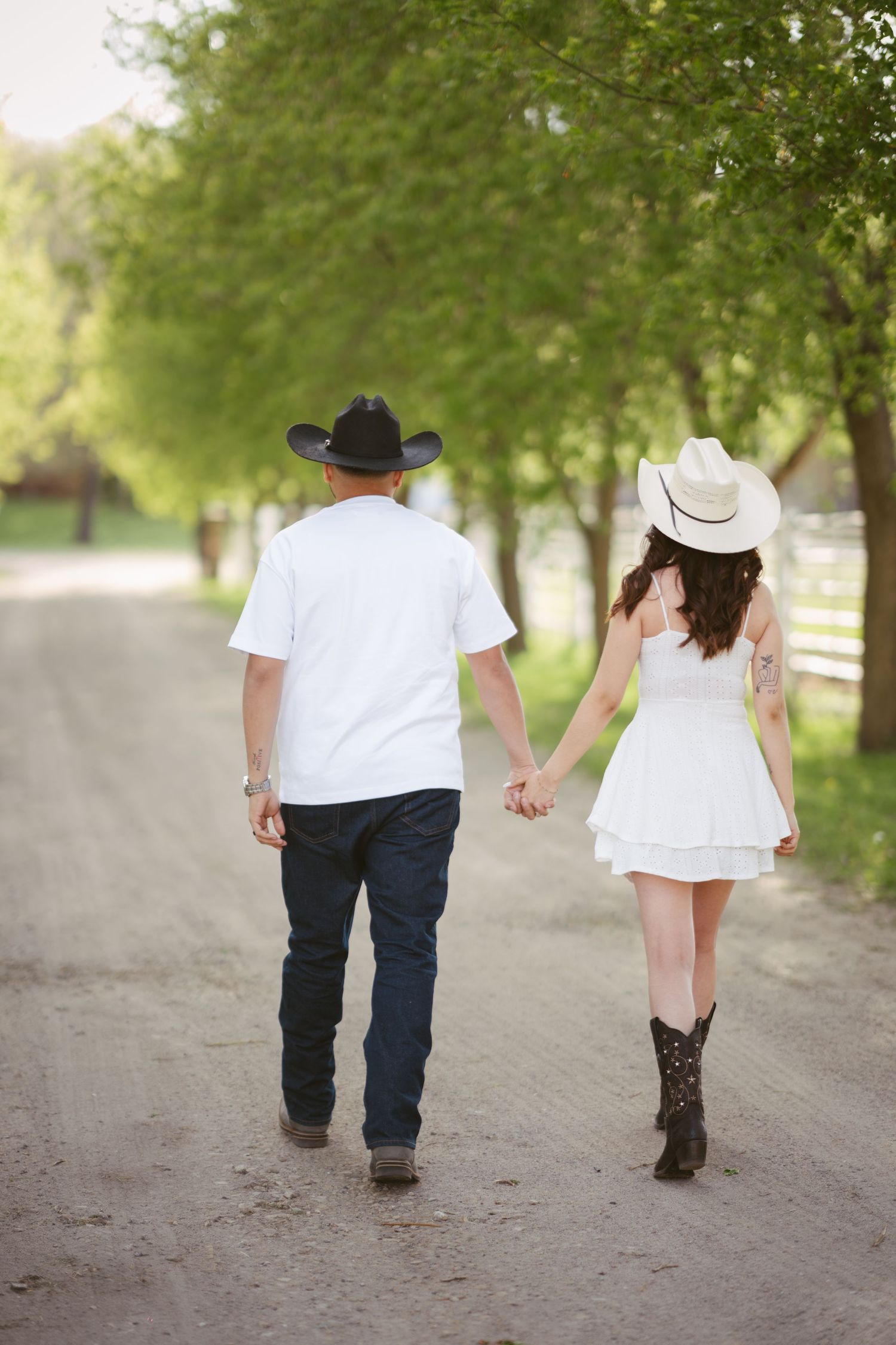 A couple in cowboy hats and white outfits walks hand-in-hand down a tree-lined dirt path.