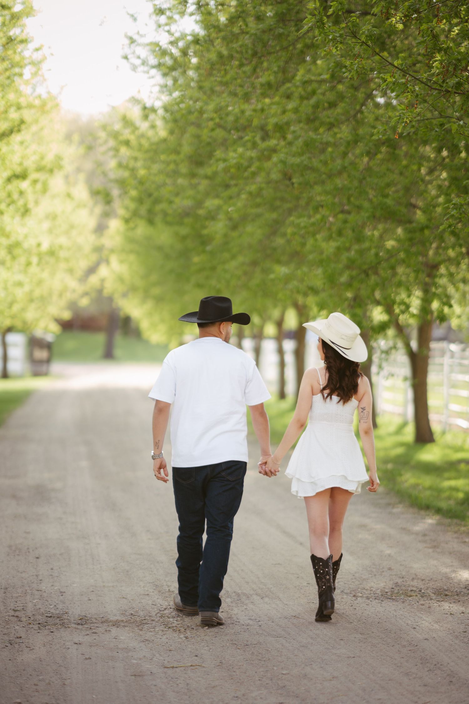 A couple in cowboy hats walks hand-in-hand down a tree-lined dirt path, woman in white dress and boots.