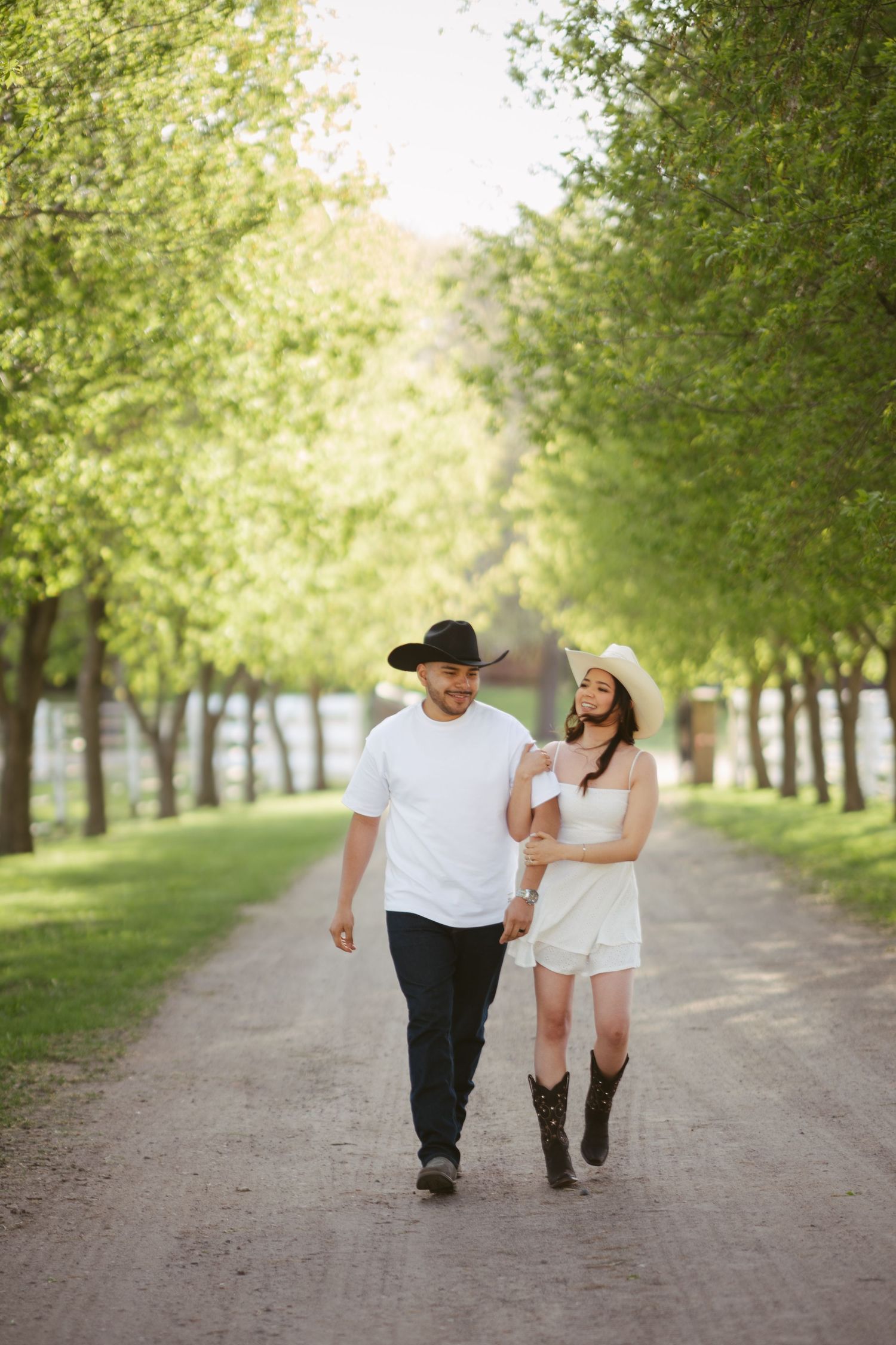 Couple in white outfits and cowboy hats walking hand in hand down a tree-lined gravel path on a sunny day.