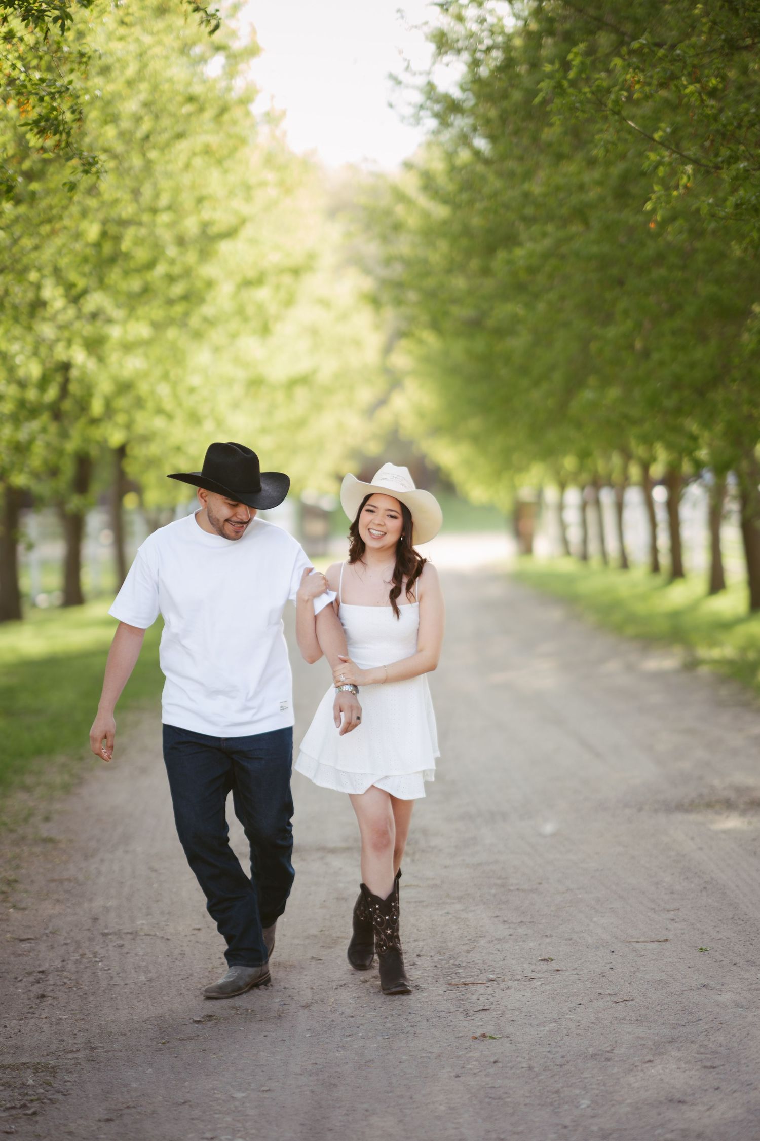 Couple walking hand in hand down a tree-lined dirt path, both wearing cowboy hats and casual western attire.