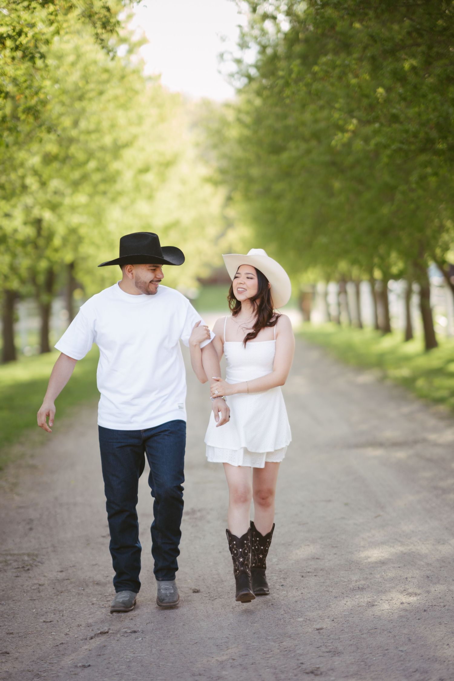 Couple in cowboy hats walking hand-in-hand down a tree-lined dirt path in matching white outfits.