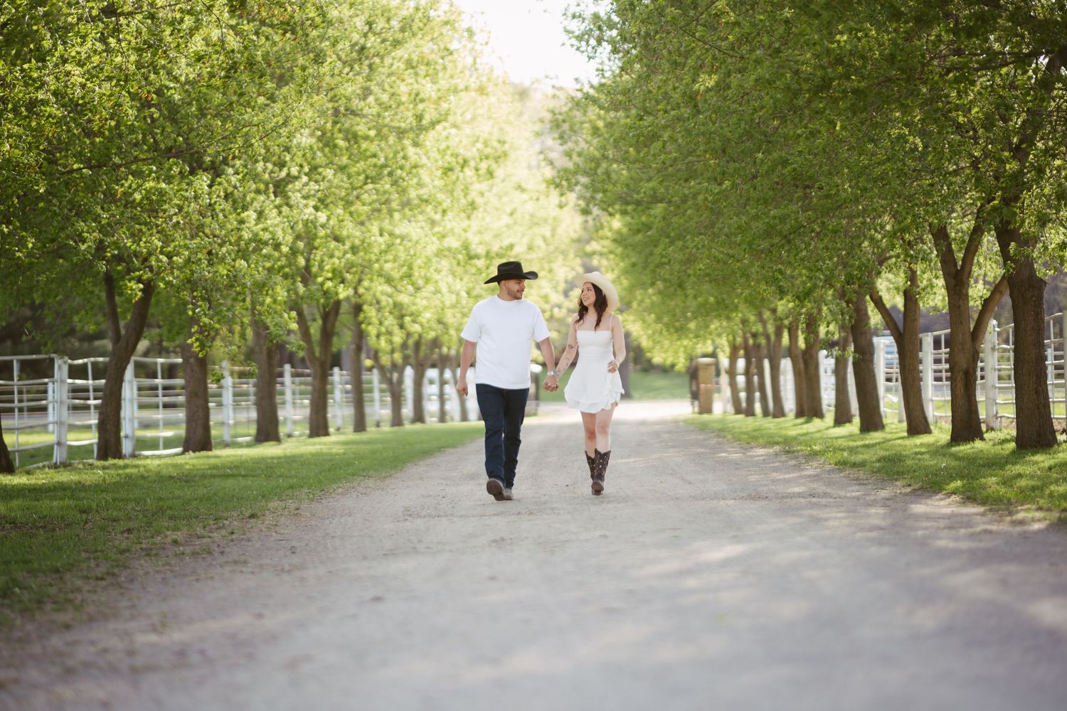 Couple in western attire walking hand-in-hand down a tree-lined path on a sunny day.